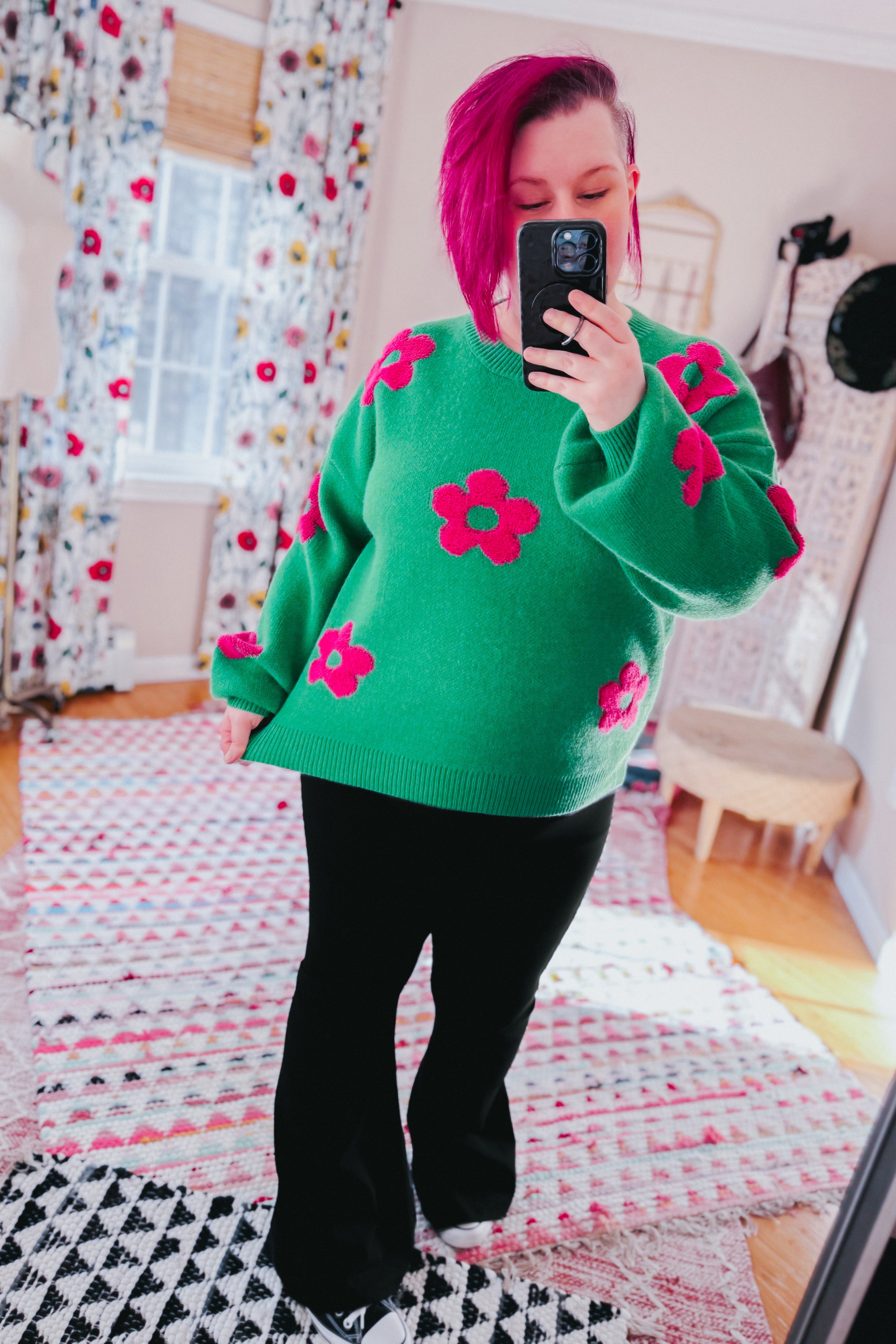 Woman wearing a green sweater embellished with tufted pink flowers and black yoga pants in an indoor setting.