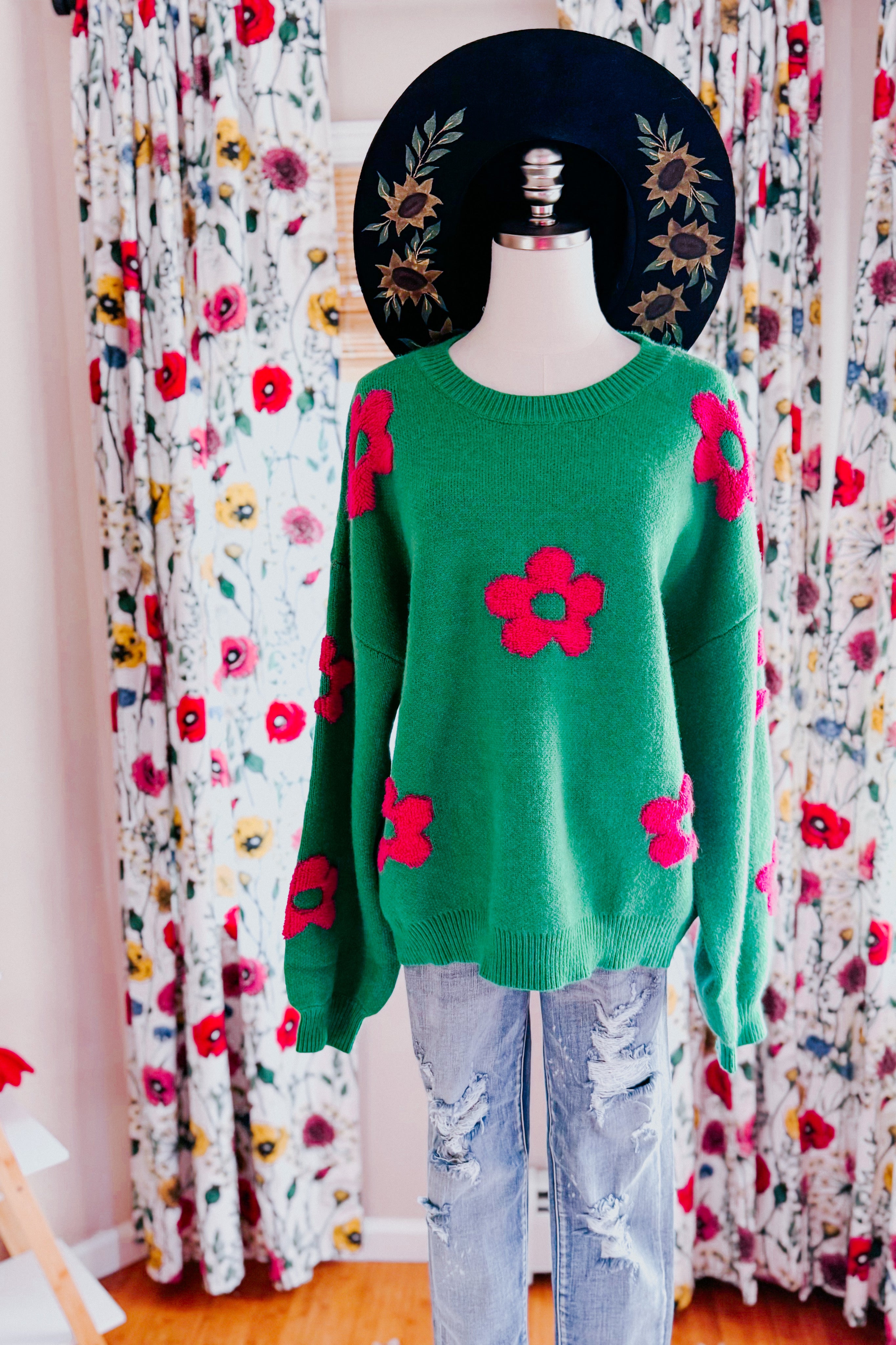 A dress form wearing a green sweater embellished with tufted pink flowers, a painted wide brim hat and blue jeans in an indoor setting.