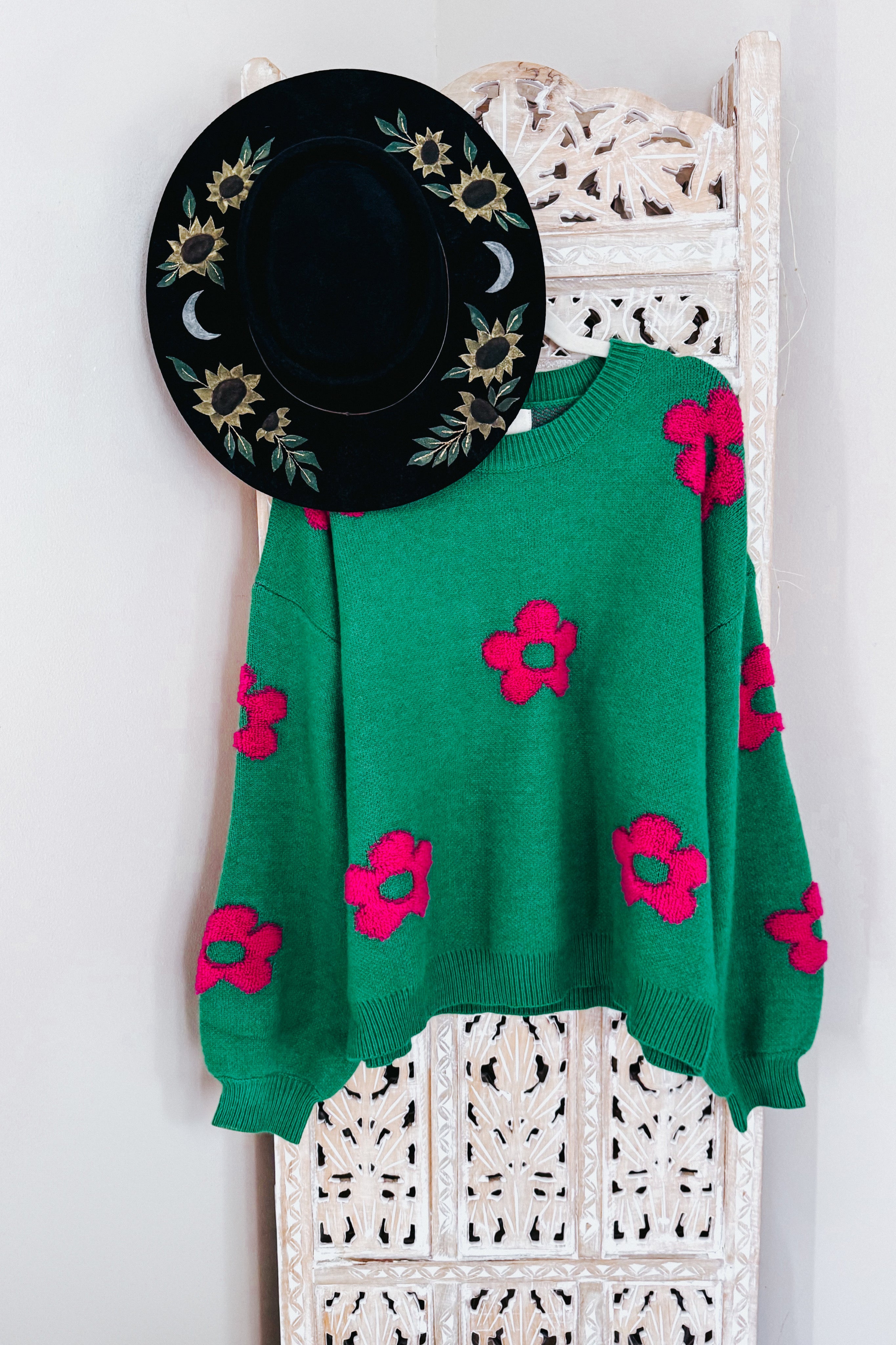 A green sweater embellished with tufted pink flowers on a hanger hanging from a rustic room divider along with a painted wide brim hat.