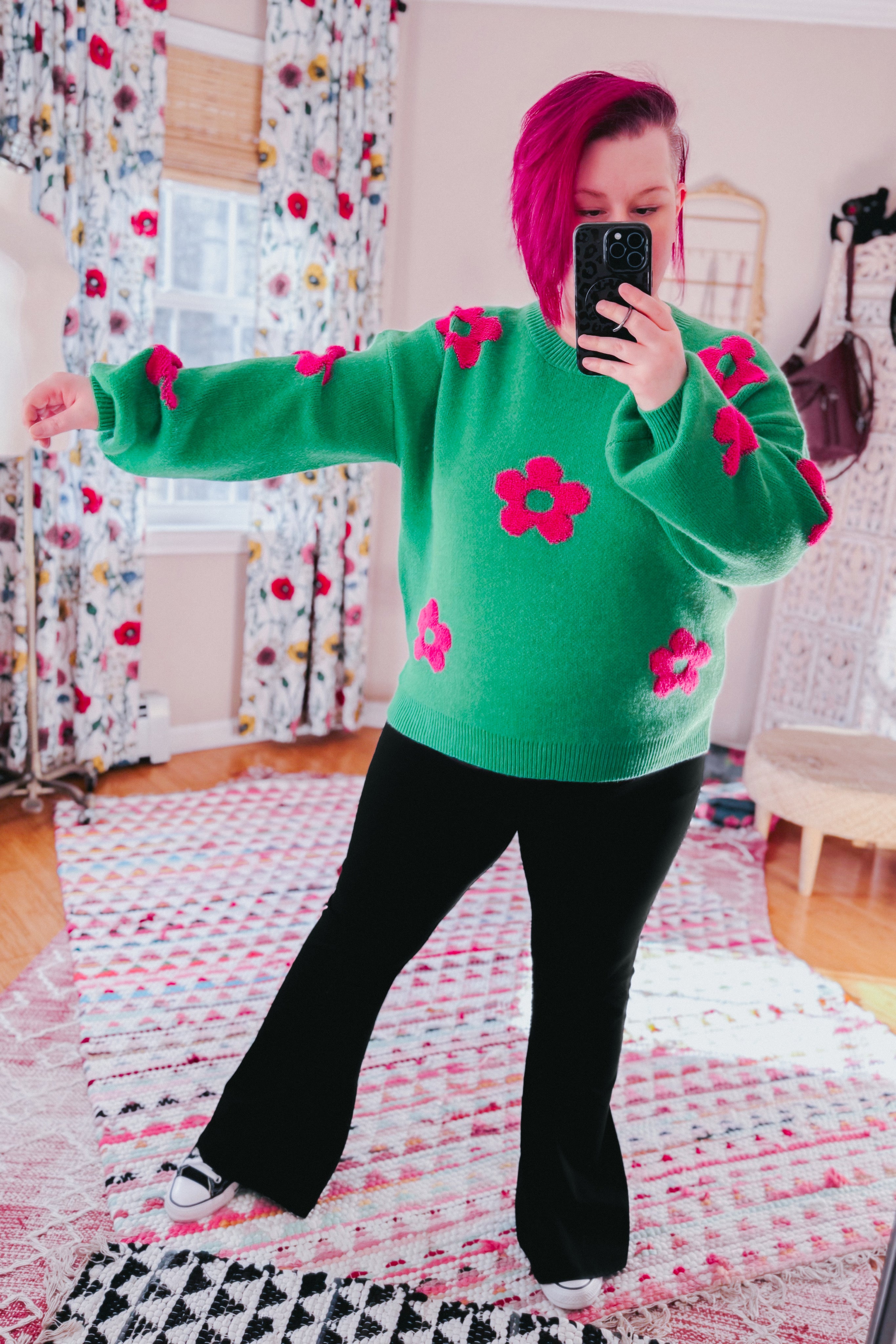 Woman wearing a green sweater embellished with tufted pink flowers and black yoga pants in an indoor setting.