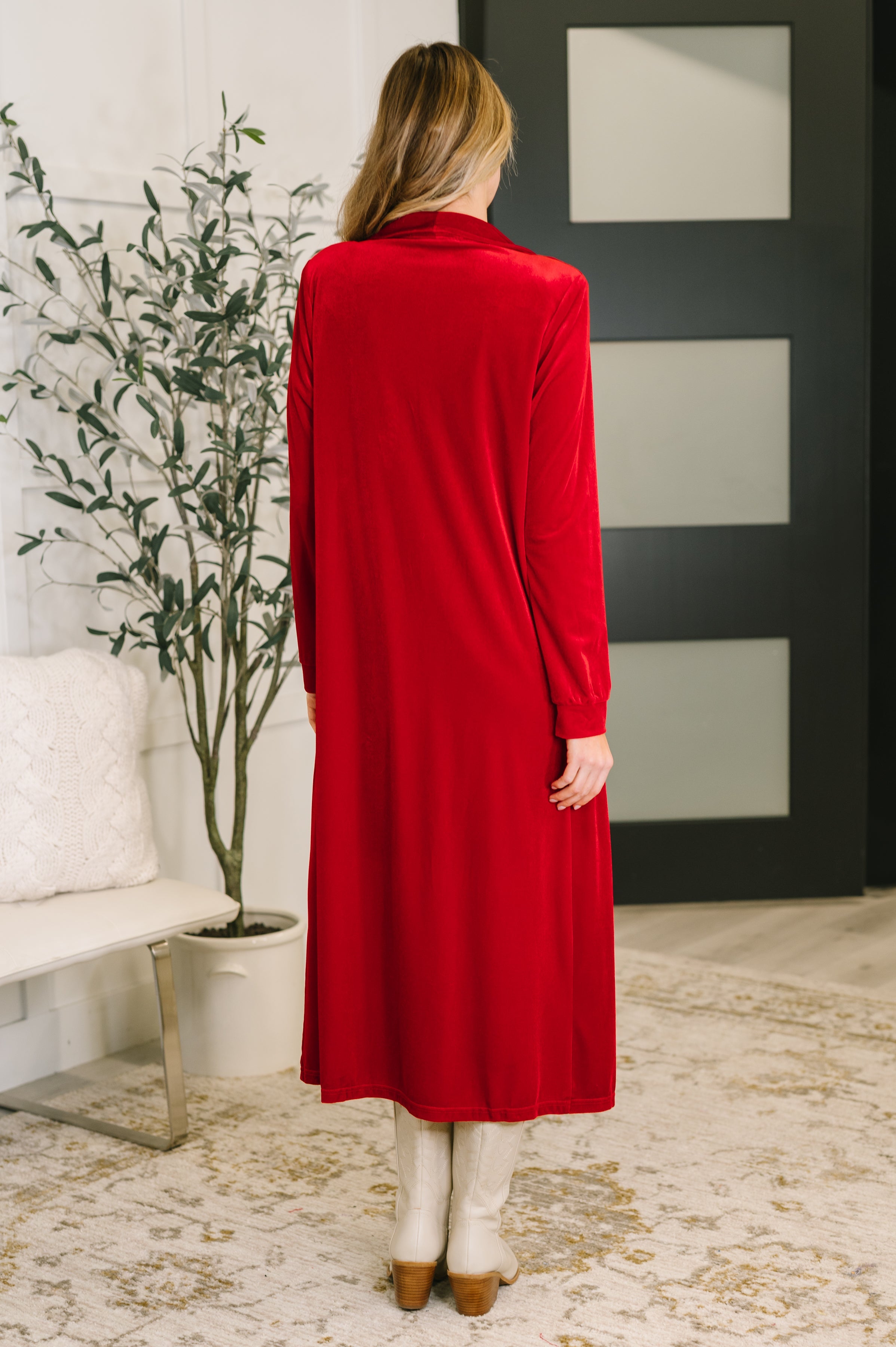 A woman wearing a red cardigan dress with her back facing the camera standing in a room with a plant and white wall.