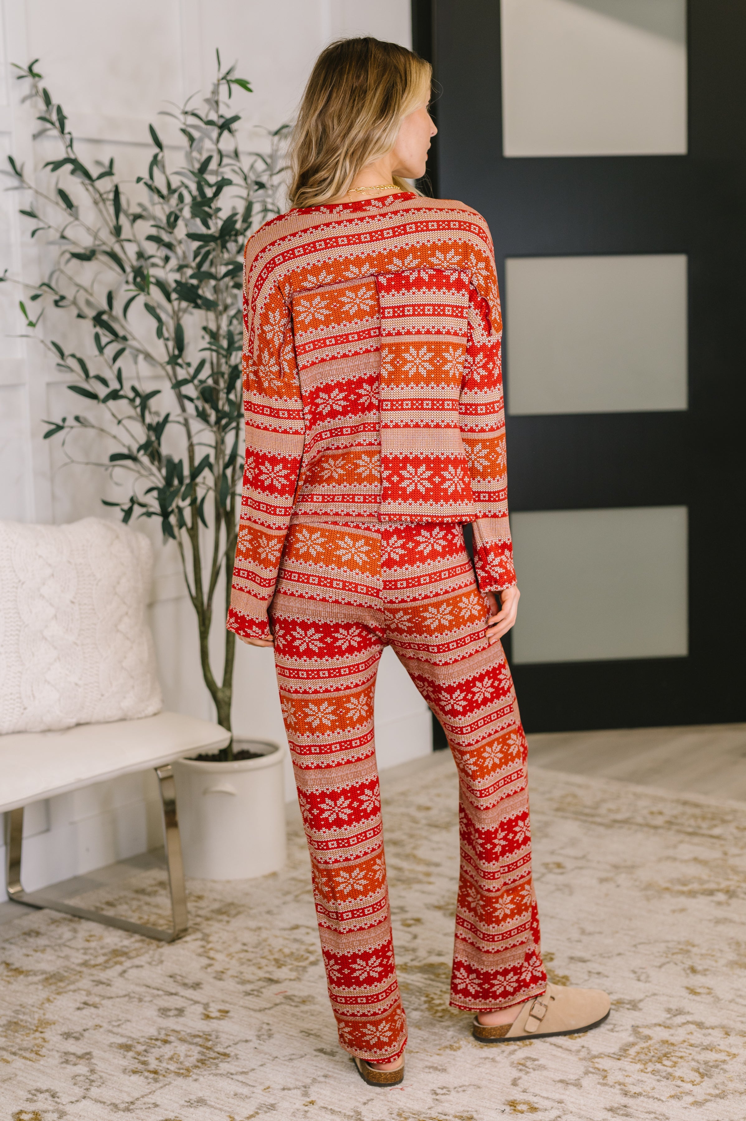 A woman with her back facing the camera wearing a red and beige snowflake patterned pajama set in a room with a plant and white bench.