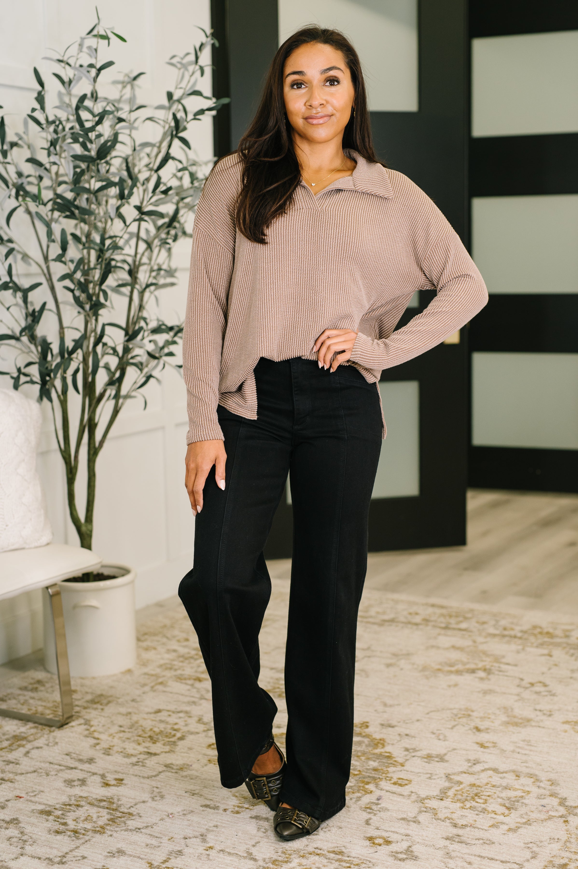 Woman wearing a beige ribbed henley collared pullover top, black jeans and black heels standing in an indoor setting.
