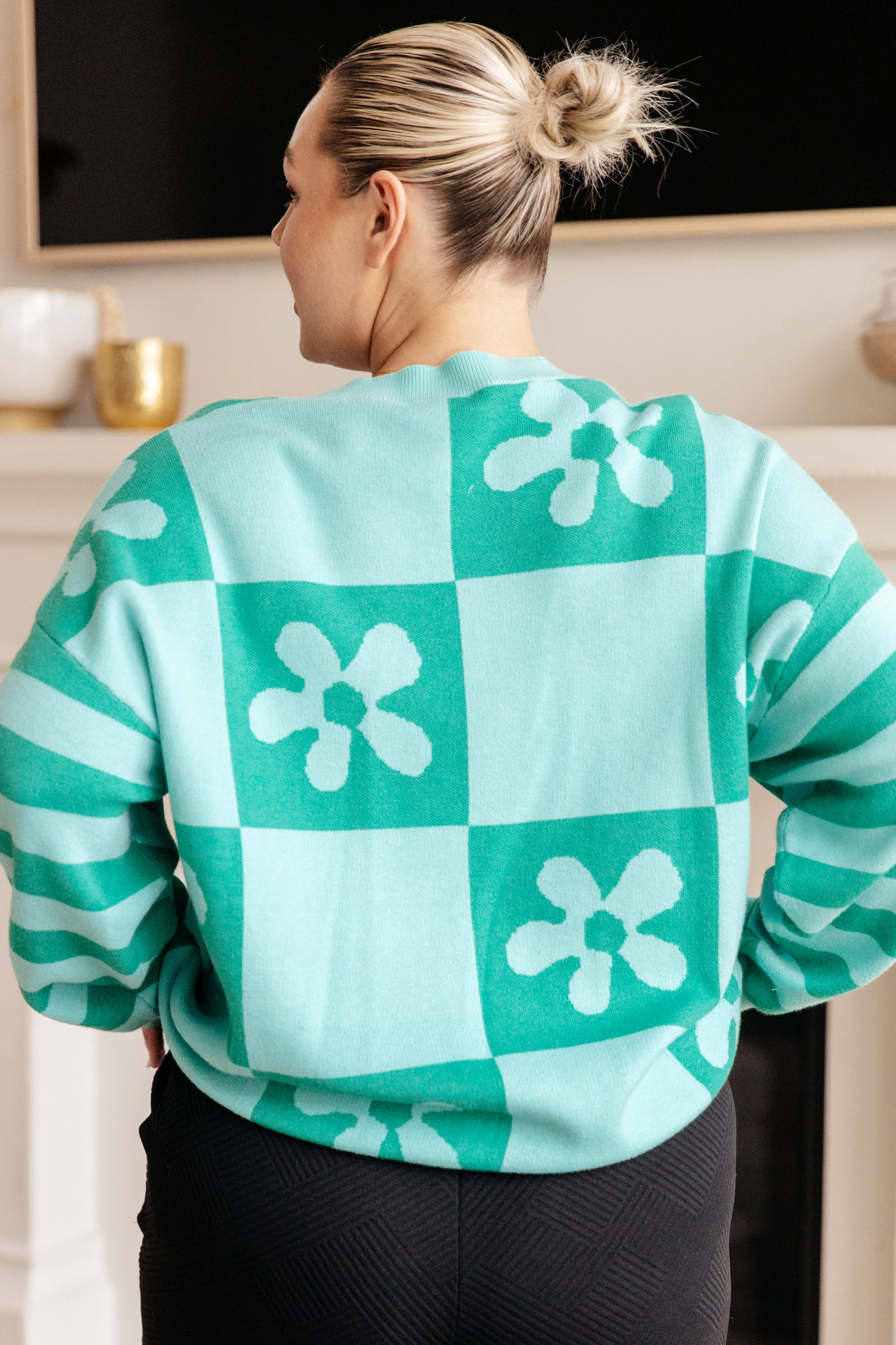Woman wearing a mint floral checkered print sweater and black pants indoors.