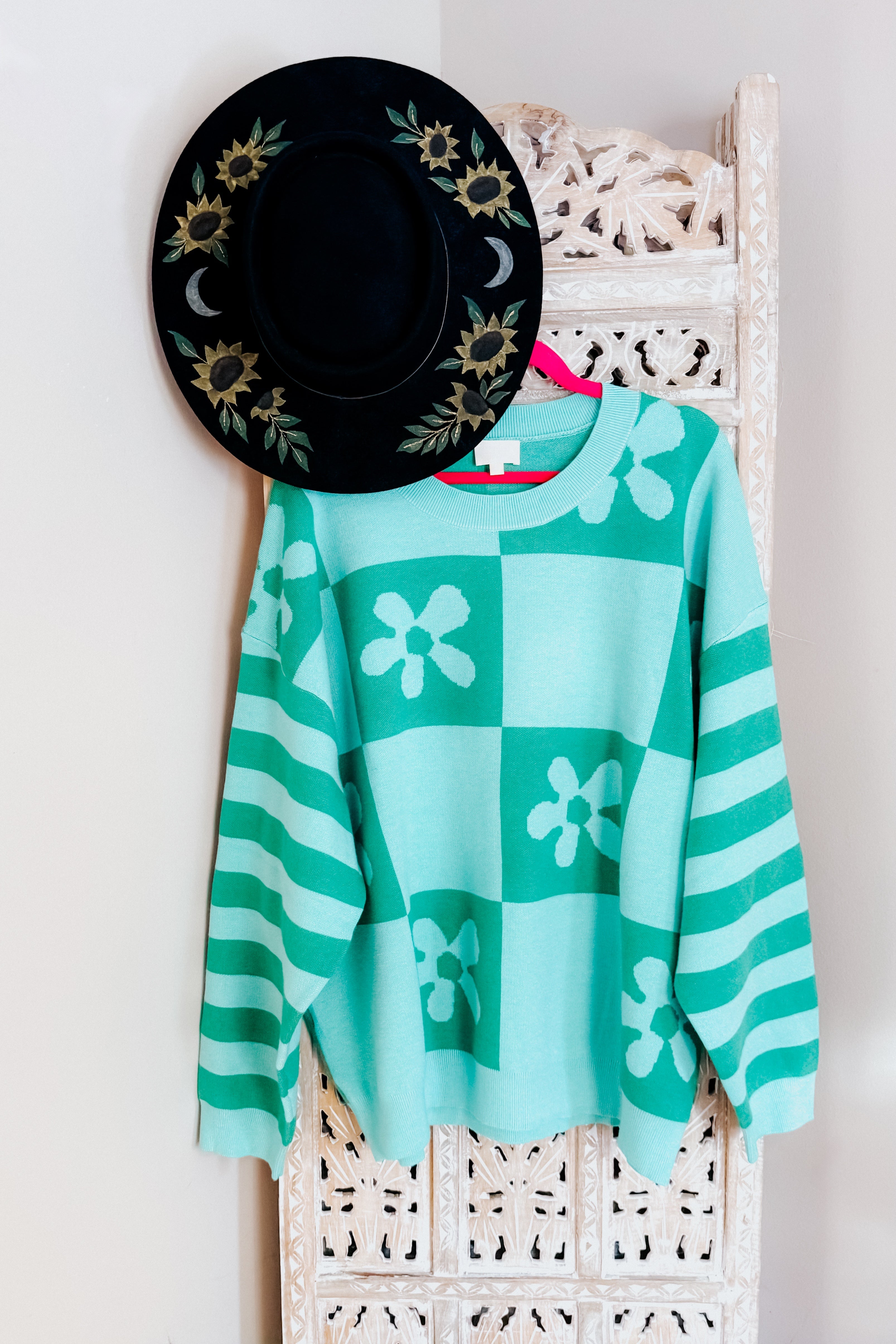 A checkered floral print mint sweater hanging from a rustic room divider with a painted wide brim hat.