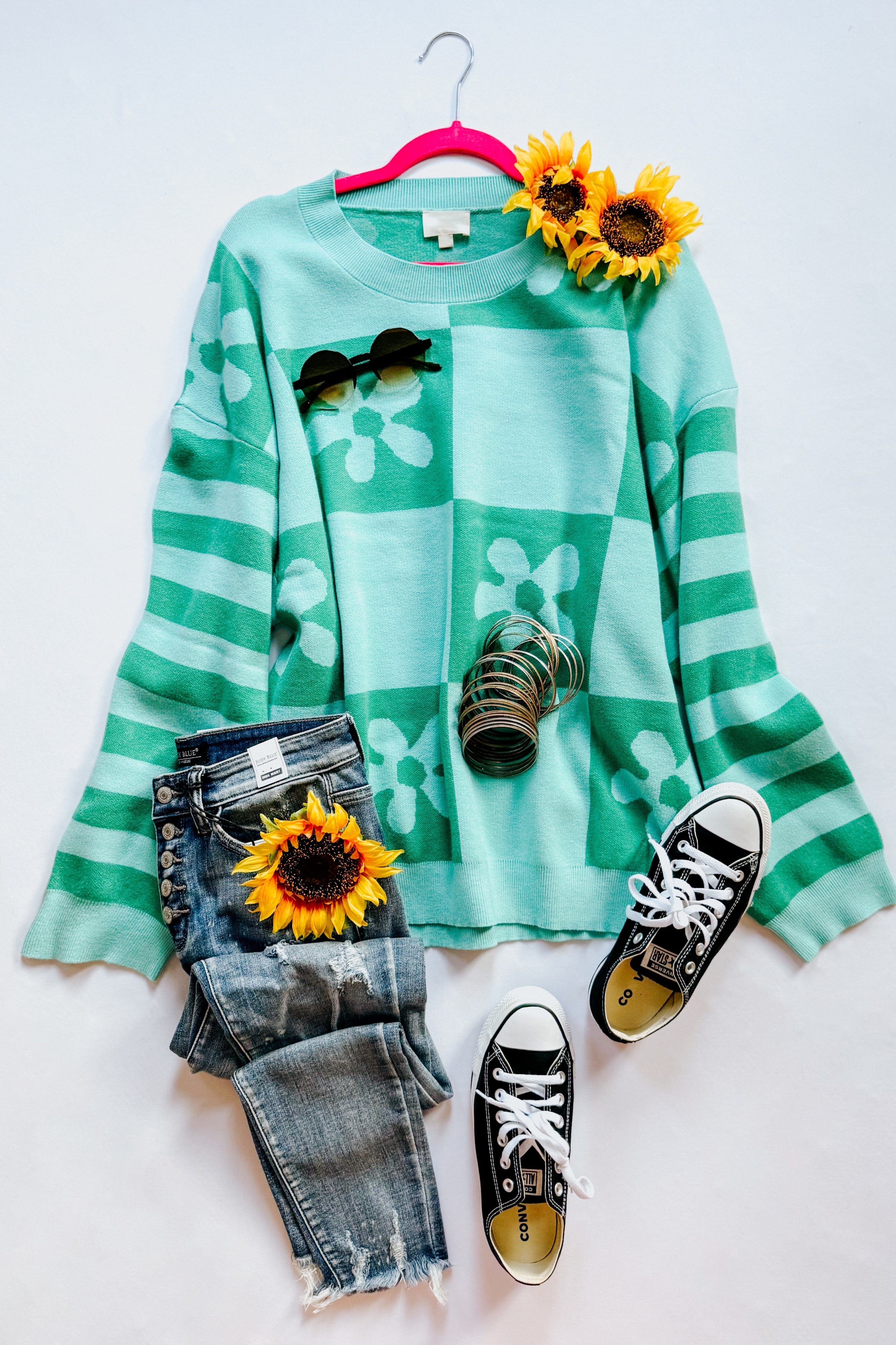 A flat lay setting of a mint floral checkered print sweater, blue jeans, black converse sneakers, accessories and flowers on a white background.