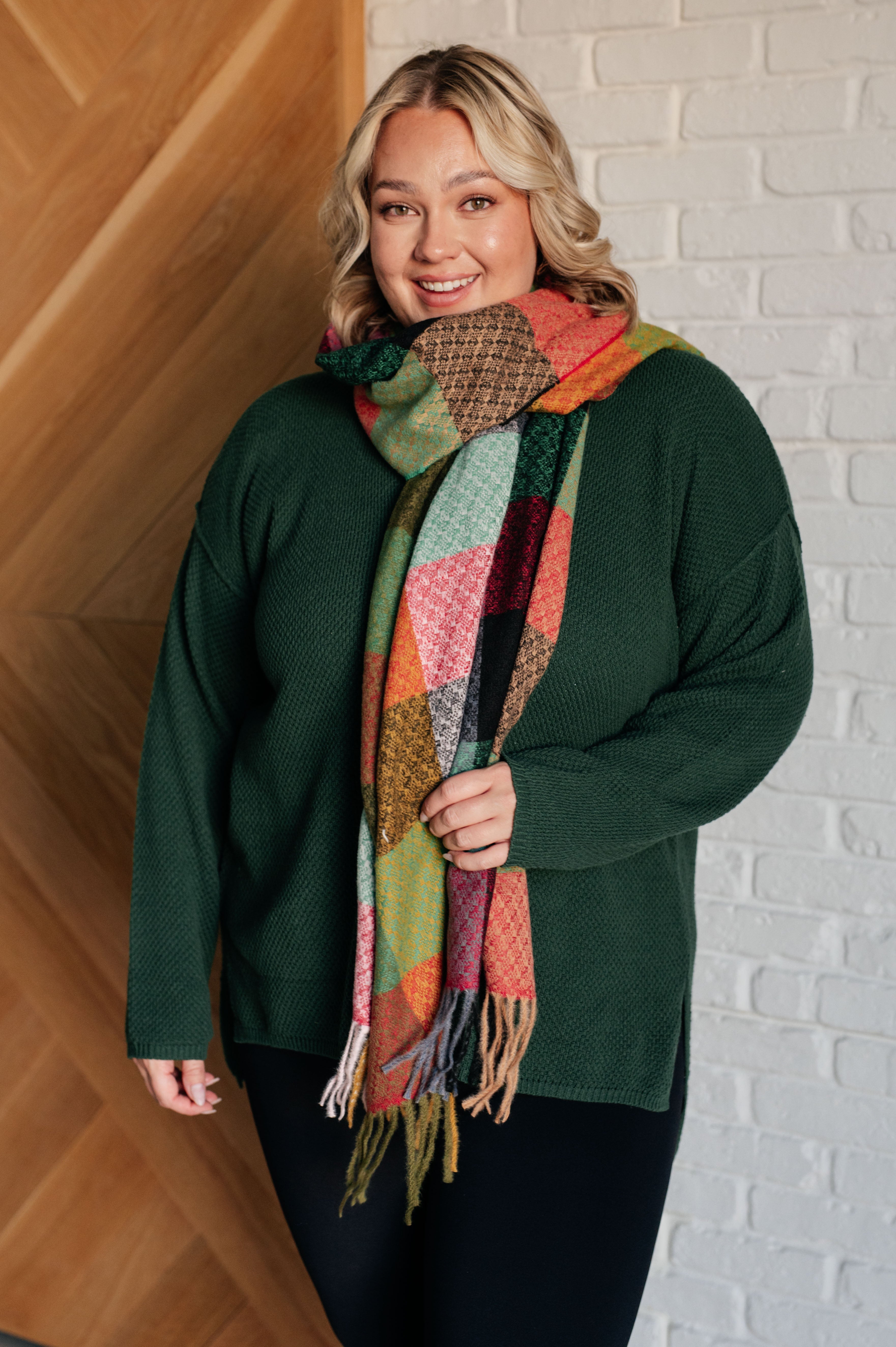 Woman wearing a green sweater, black plants and a colorful checkered scarf with fringe against a white brick wall.