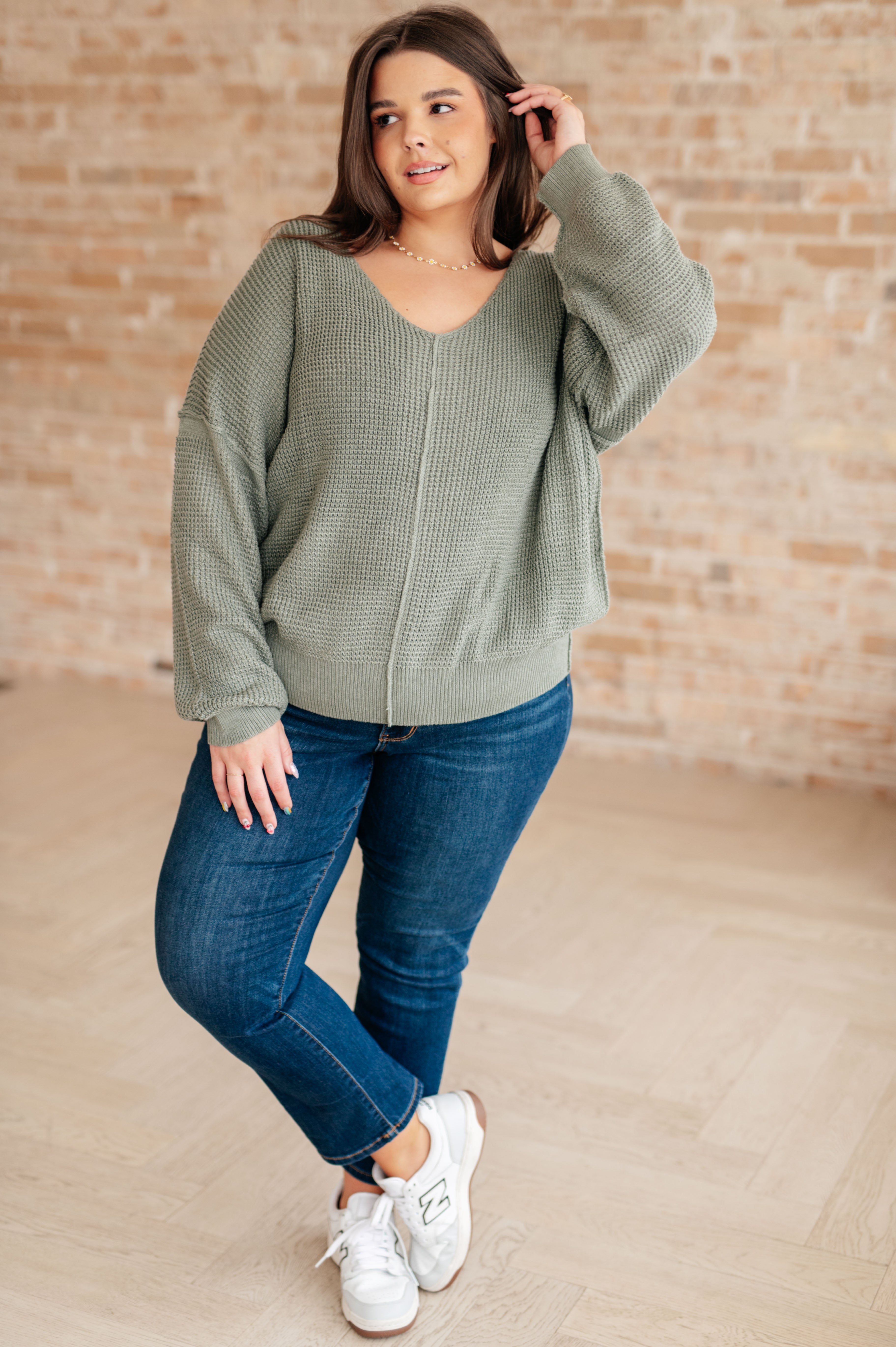 Woman wearing an olive green v-neck sweater with exposed seams, blue jeans and white sneakers standing against a brick wall