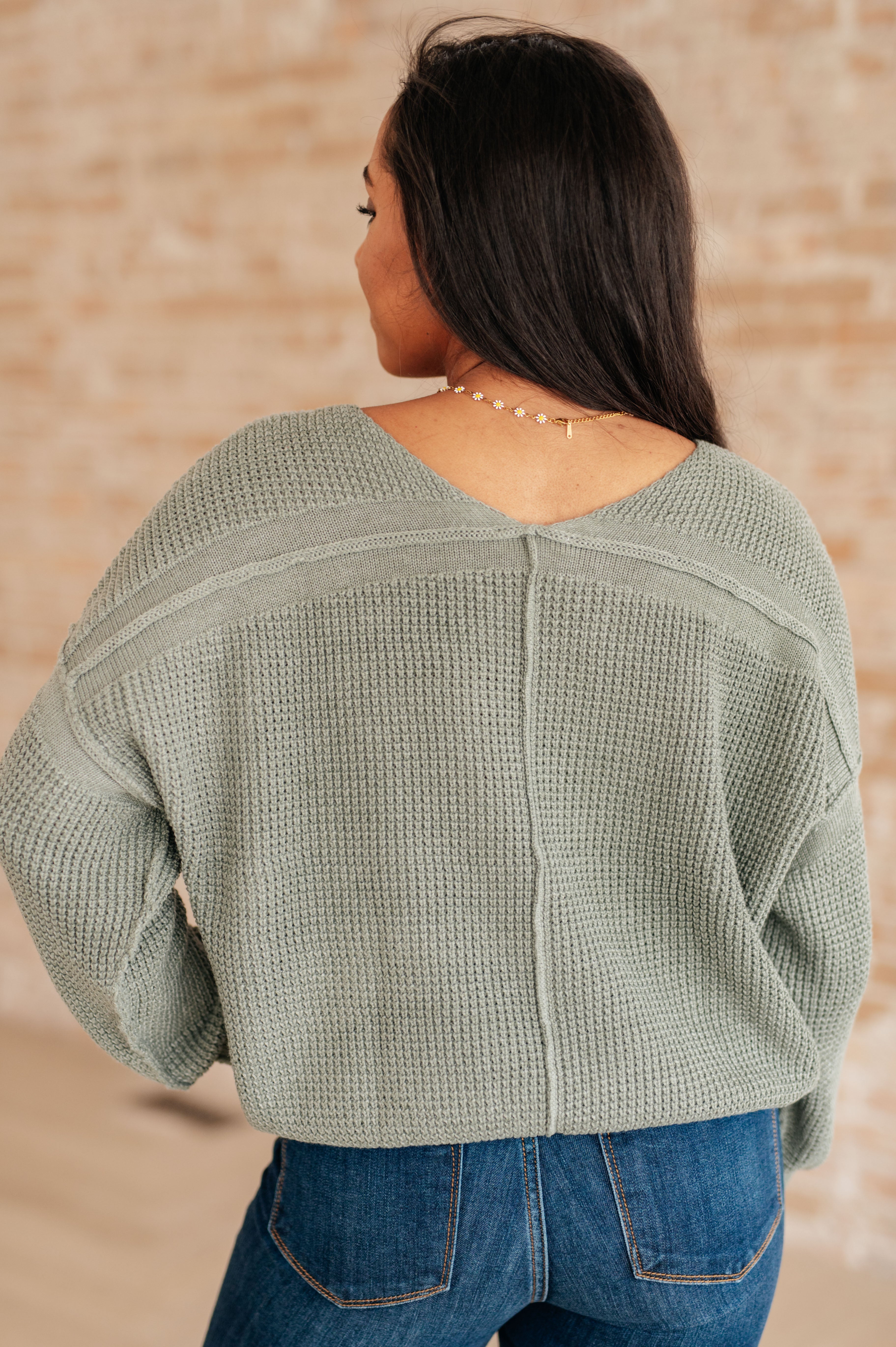 Woman wearing an olive green v-neck sweater with exposed seams and blue jeans standing against a brick wall