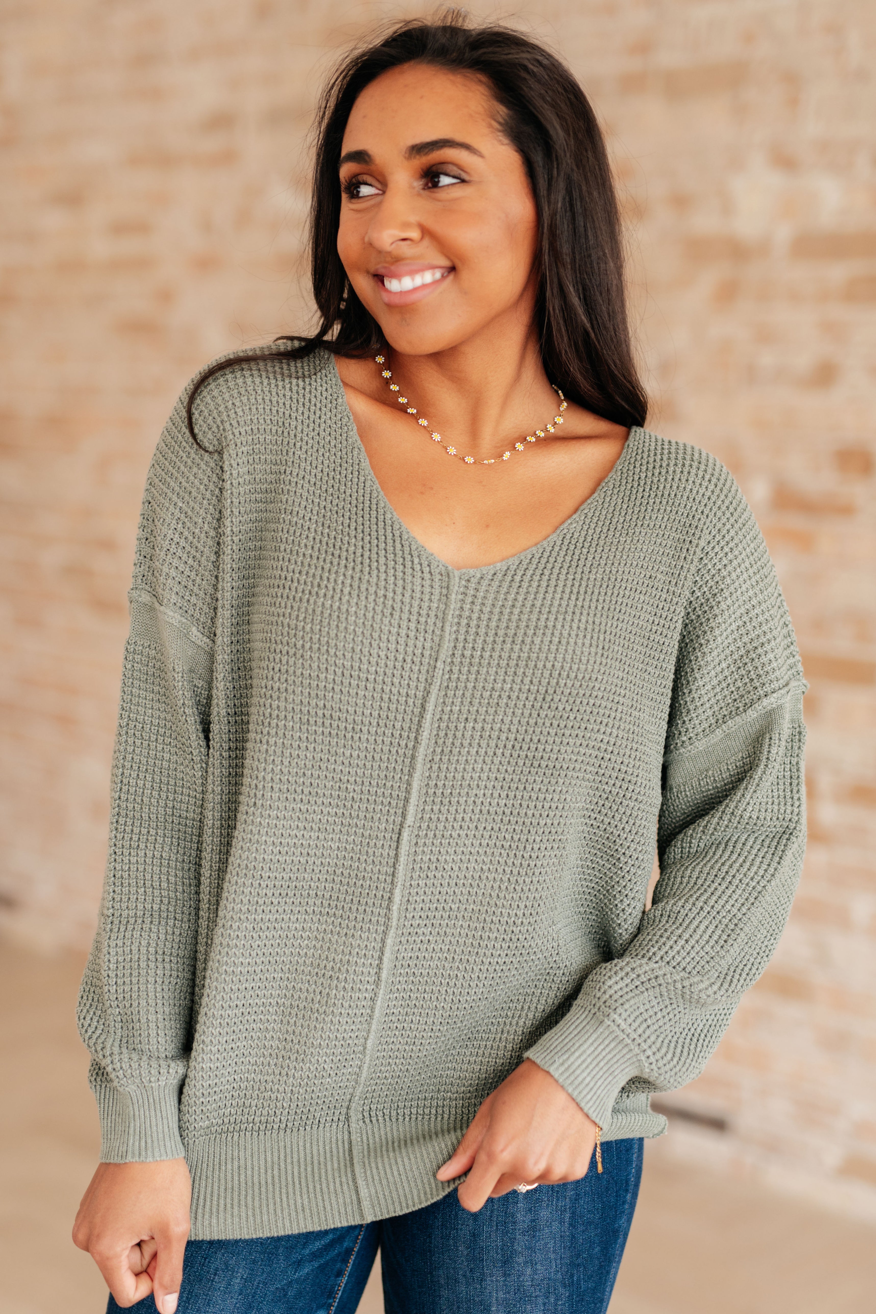Woman wearing an olive green v-neck sweater with exposed seams and blue jeans standing against a brick wall