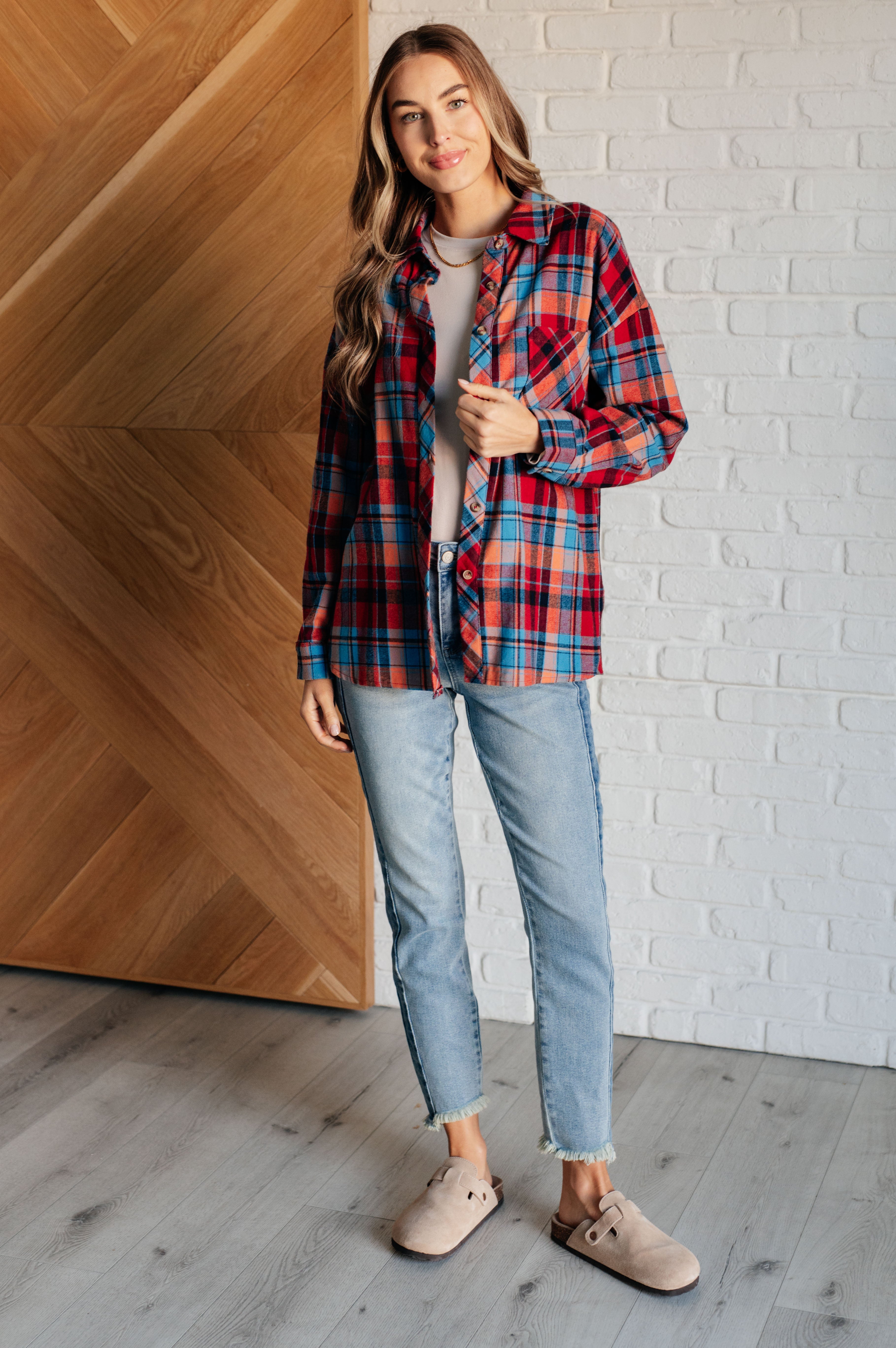 Woman wearing a red and blue long-sleeves plaid button-up shirt, a beige top, blue jeans and tan clogs with a white brick wall background.