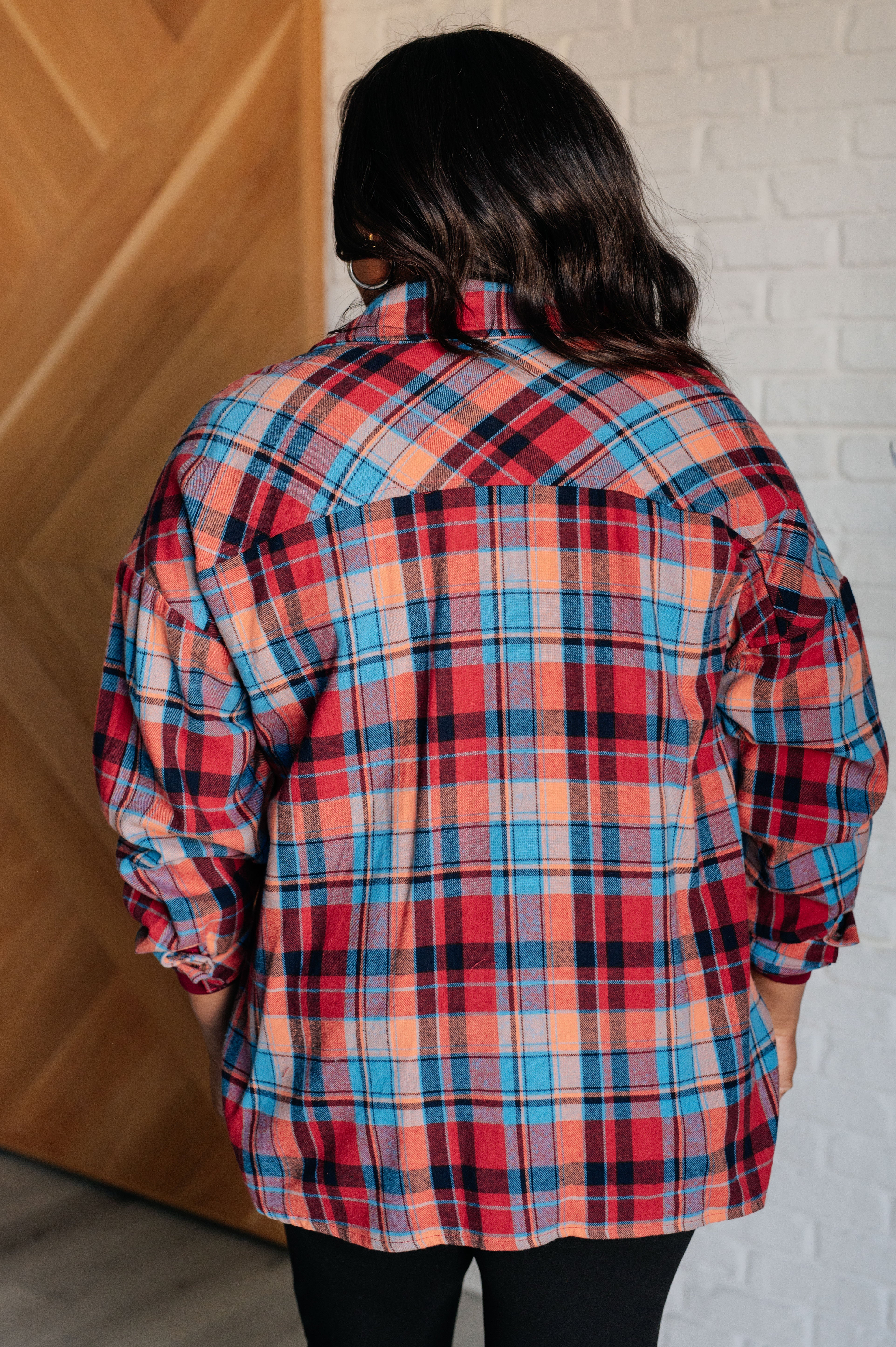 Woman wearing a red and blue long-sleeves plaid button-up shirt and black pants with a white brick wall background.