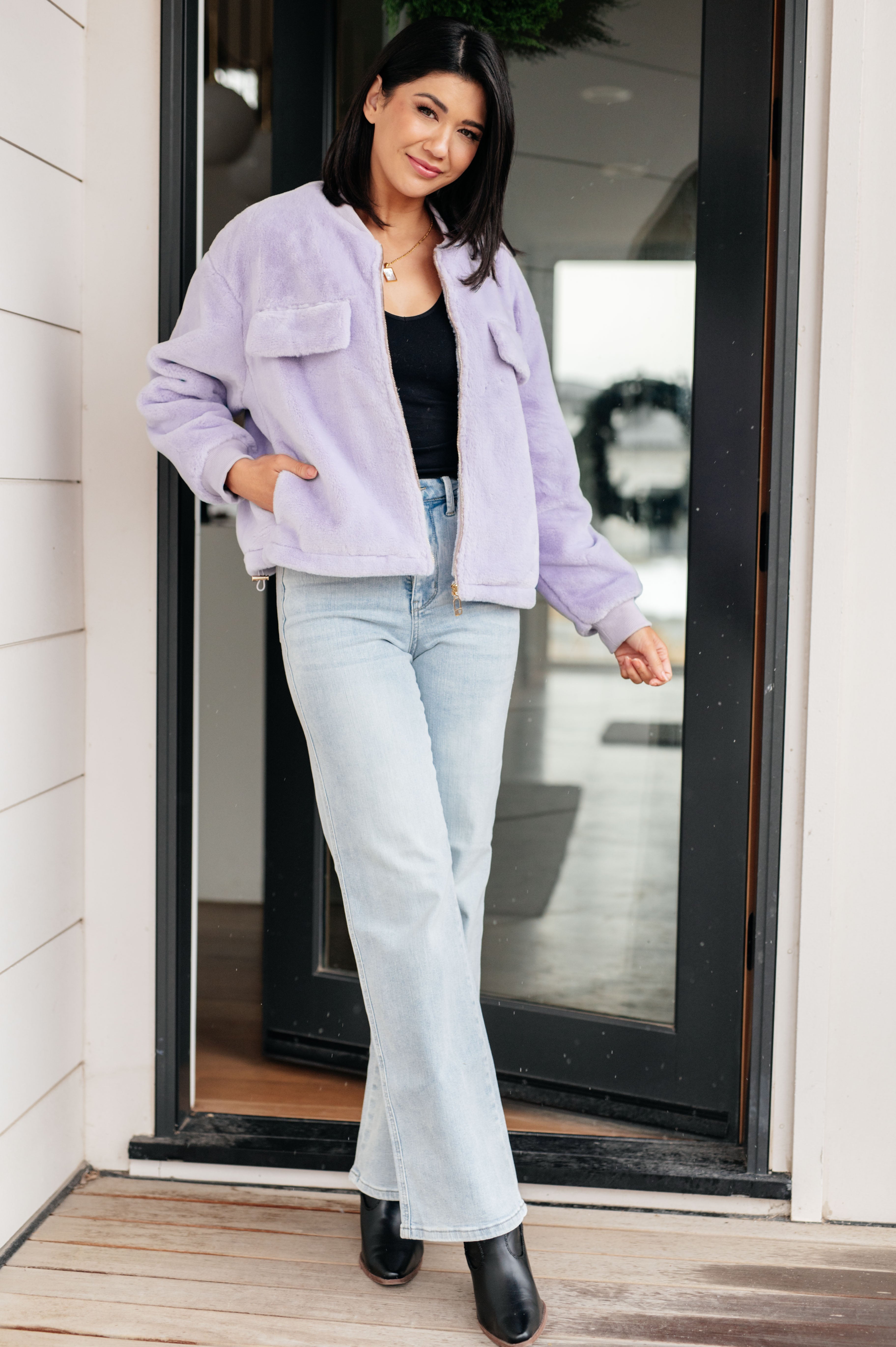 Woman wearing a light purple fuzzy zip up cropped coat, black top, light blue jeans and black boots standing in front of a glass door.