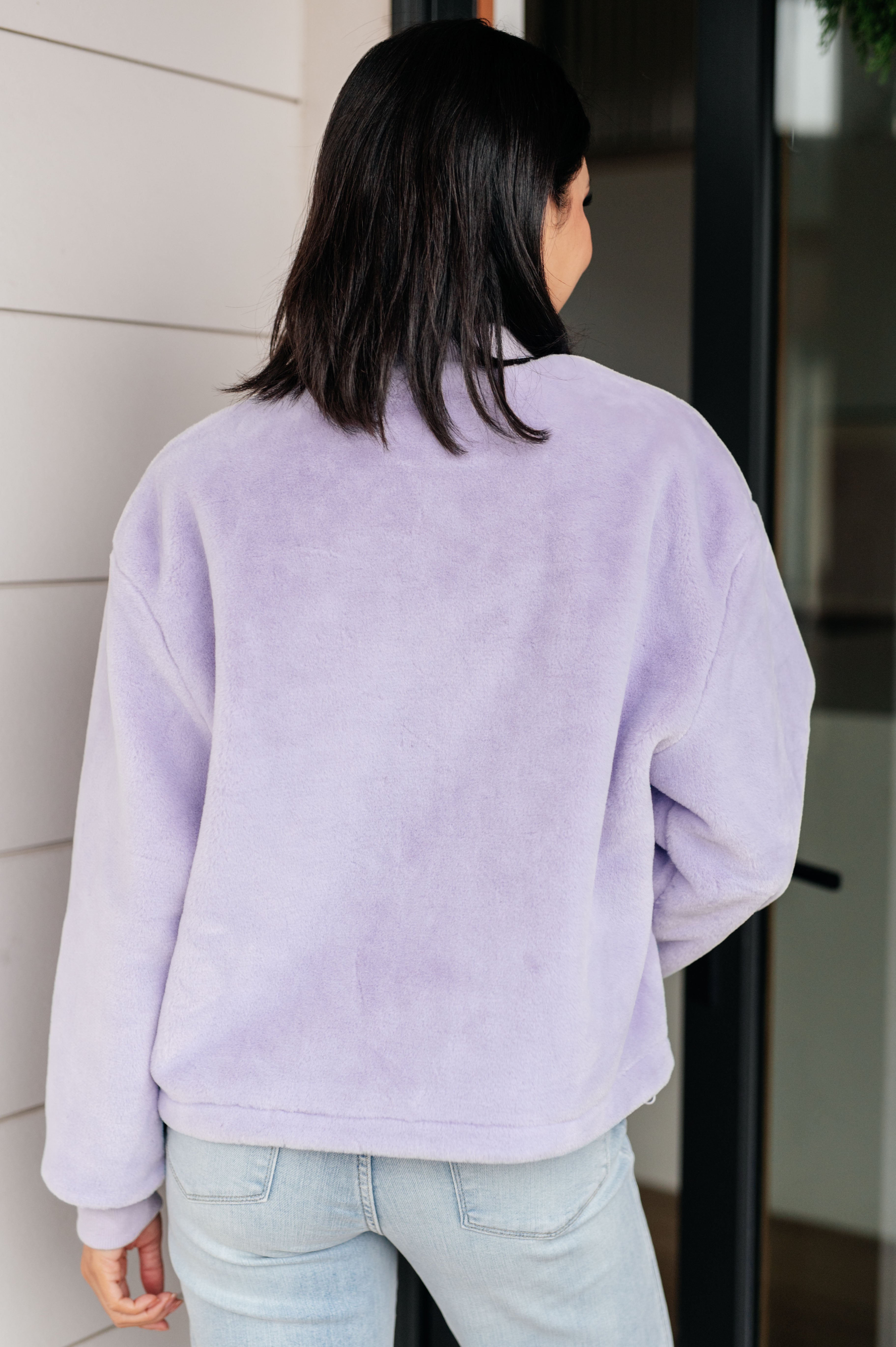 Woman wearing a light purple fuzzy zip up cropped coat and light blue jeans standing in front of a glass door.