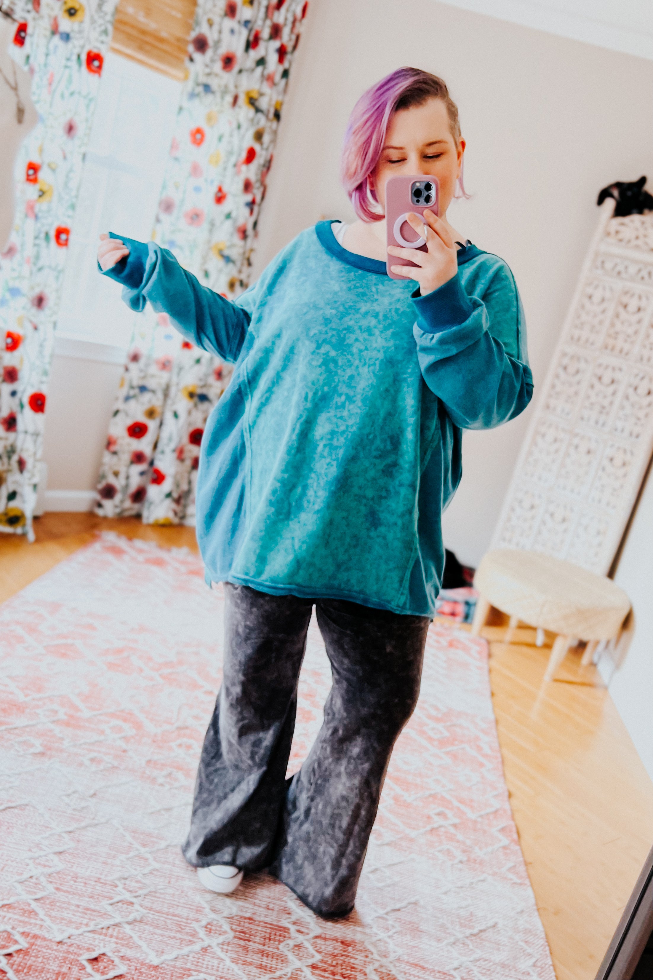 A woman with pink hair wearing a blue mineral wash sweater over dark grey flare pants against an indoor setting.
