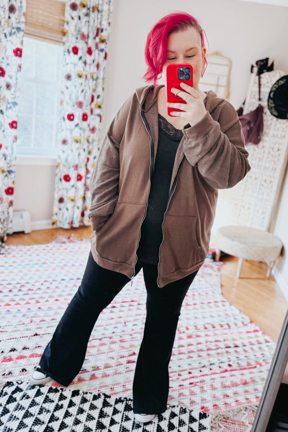 A woman with red hair wearing a brown zip up hoodie, a grey lace top and black flare pants against an indoor setting.