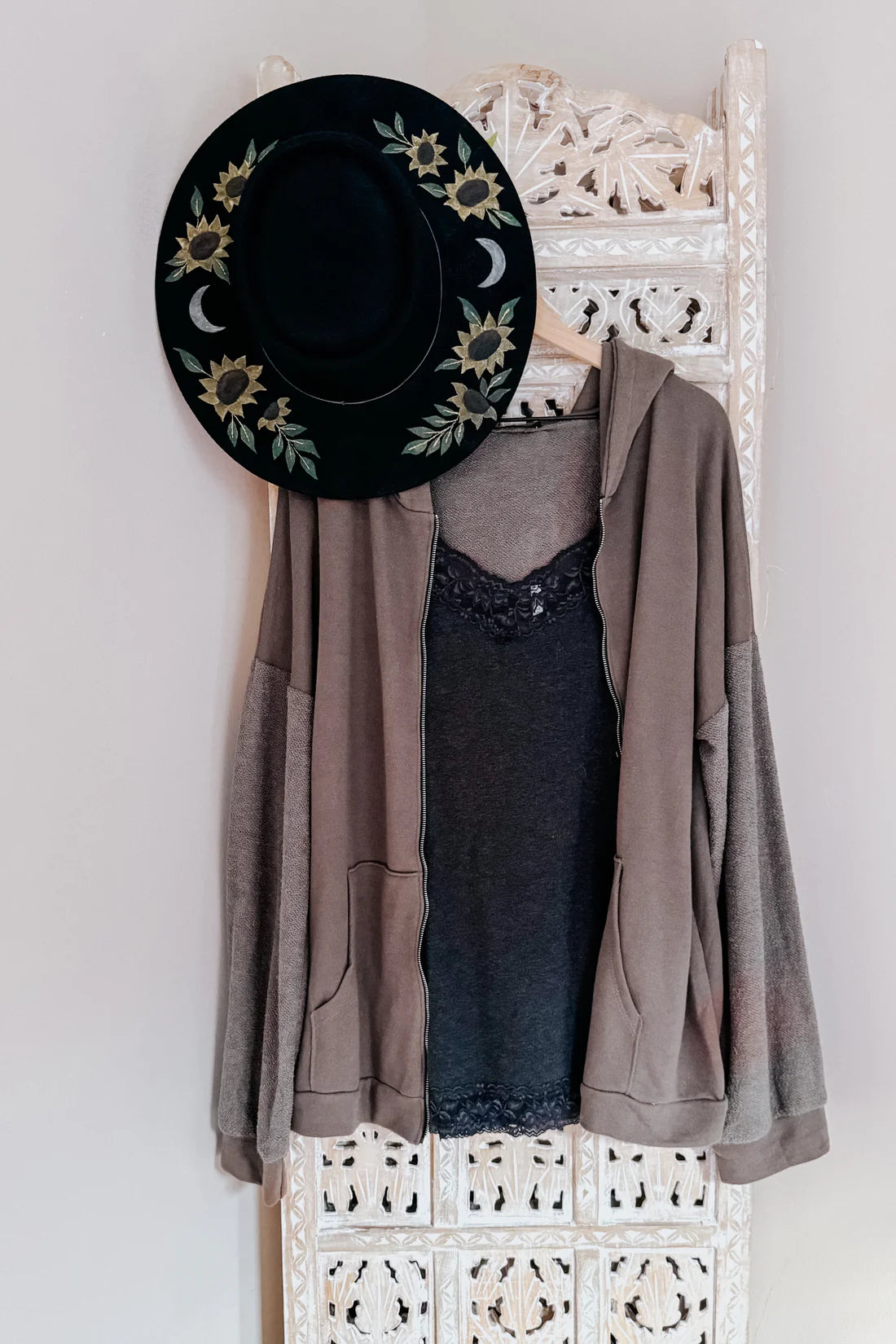 Black sun hat with floral painted accents, brown zip up hoodie, grey lace top on a white lattice rack.
