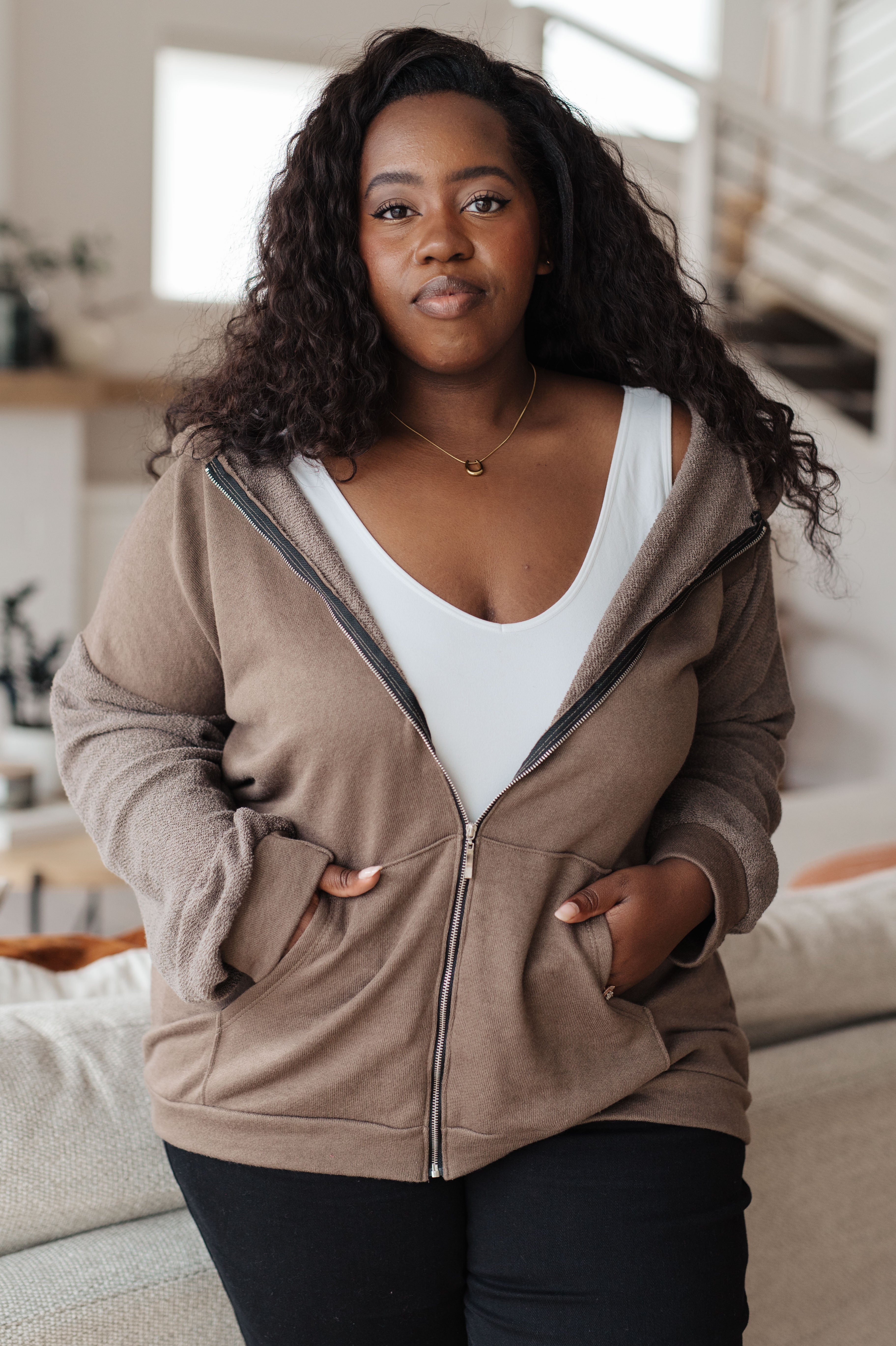 Woman wearing a brown zip-up hoodie over a white tank top over black jeans in a casual indoor setting