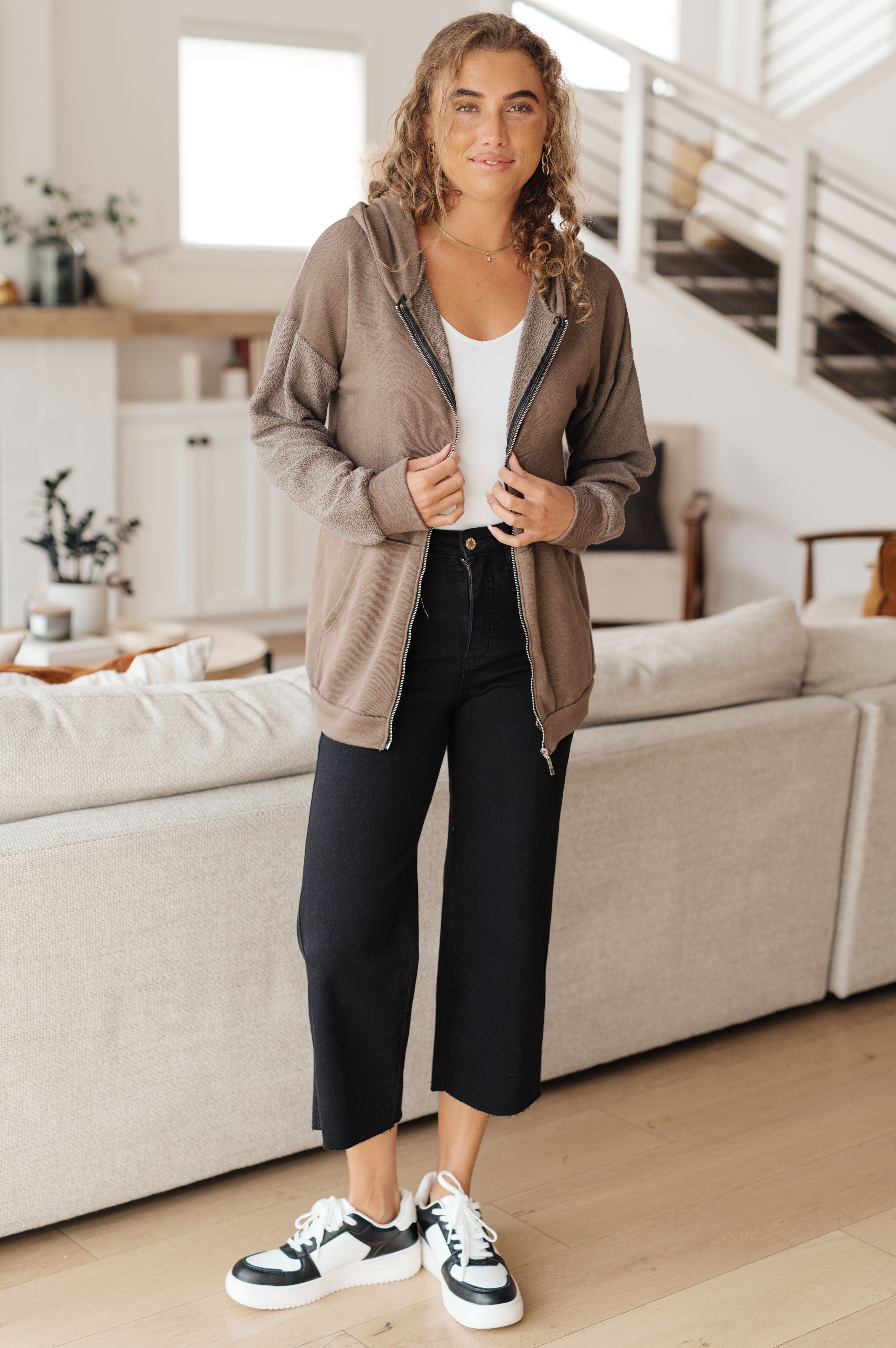 Woman wearing a brown zip-up hoodie over a white tank top over black jeans and black and white sneakers in a casual indoor setting.