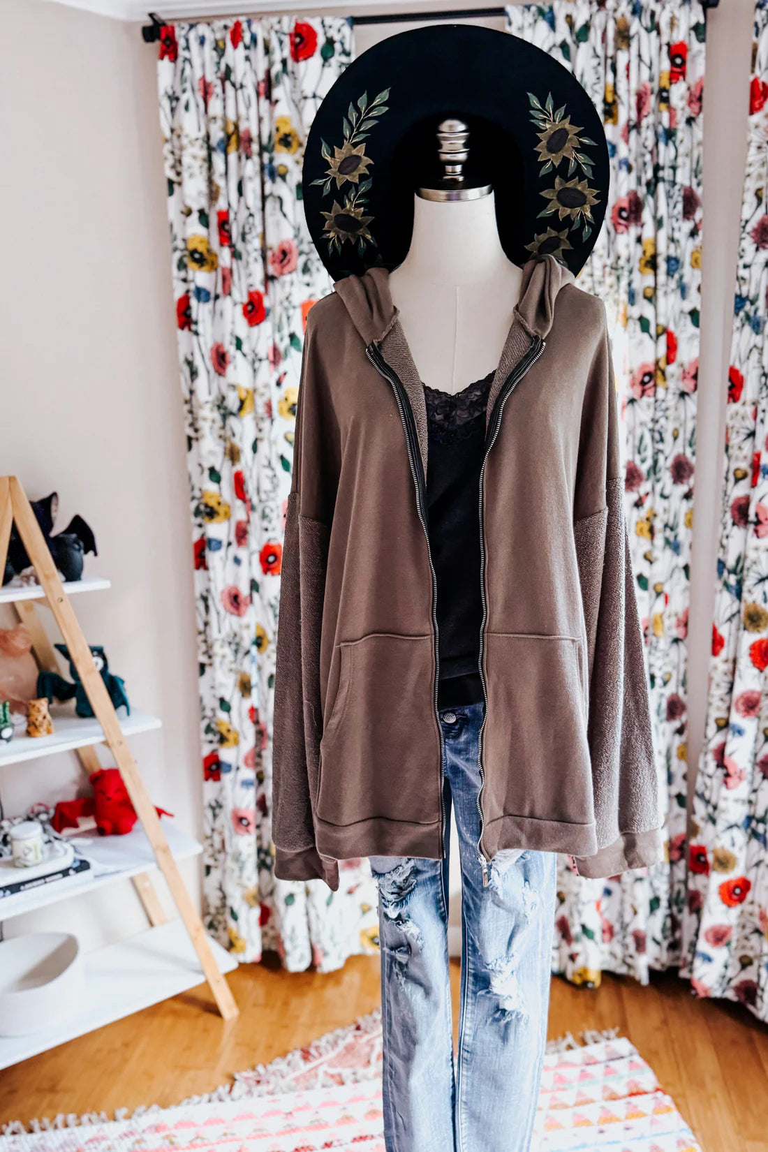 A dress form wearing a black sunflower hat, a grey lace top with a brown zip up hoodie over blue jeans against an indoor setting.