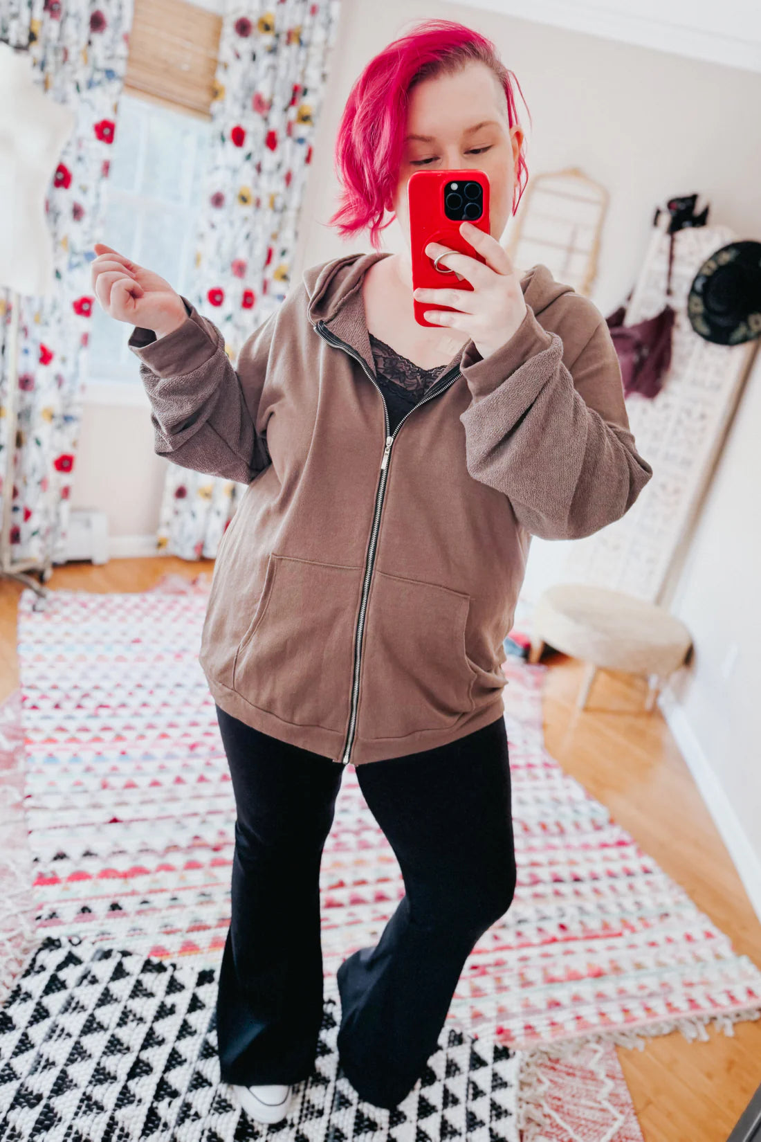 A woman with red hair wearing a brown zip up hoodie, a grey lace top and black flare pants against an indoor setting.