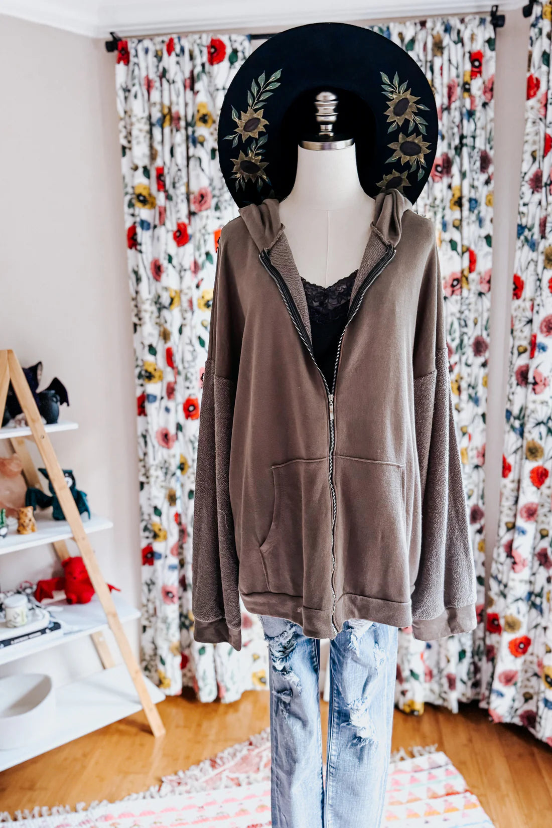 A dress form wearing a black sunflower hat, a grey lace top with a brown zip up hoodie over blue jeans against an indoor setting.