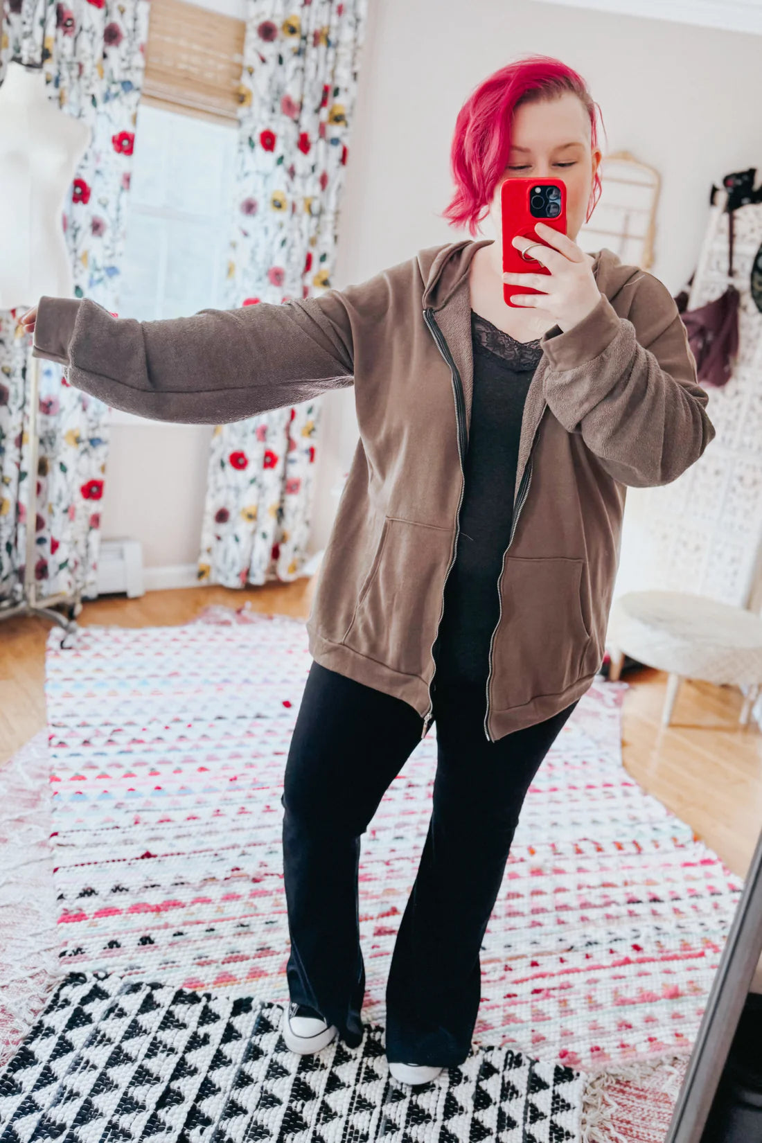 A woman with red hair wearing a brown zip up hoodie, a grey lace top and black flare pants against an indoor setting.