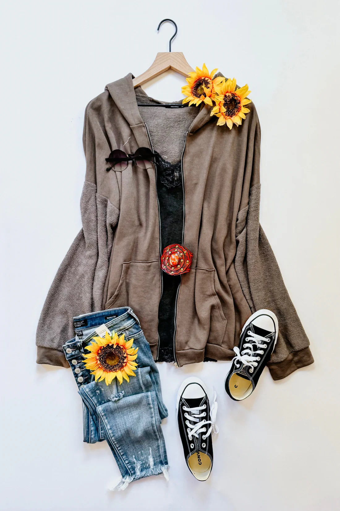 A flat lay of a brown zip up hoodie over a grey lace top, blue jeans, accessories and floral accents on a white background.