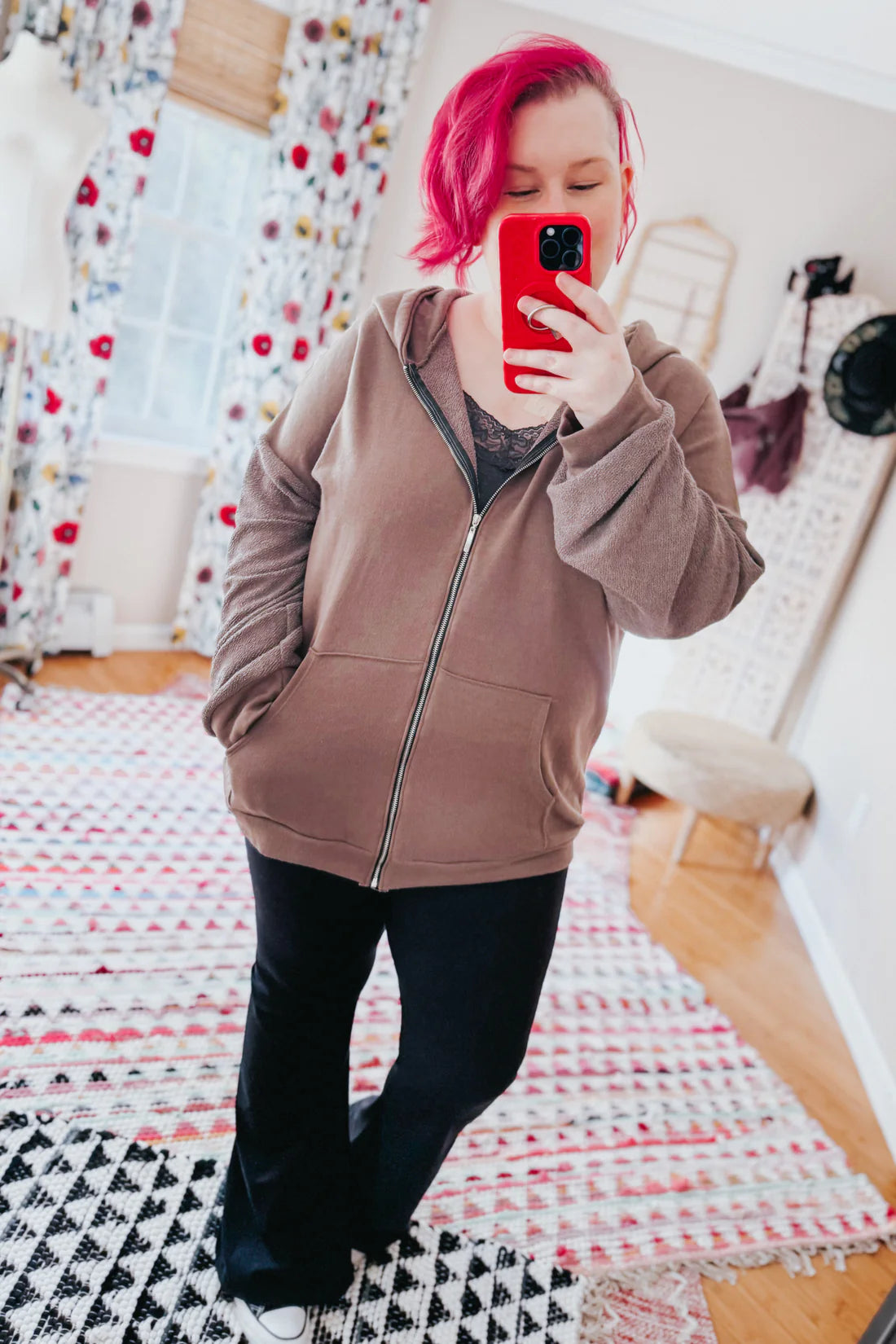 A woman with red hair wearing a brown zip up hoodie, a grey lace top and black flare pants against an indoor setting.