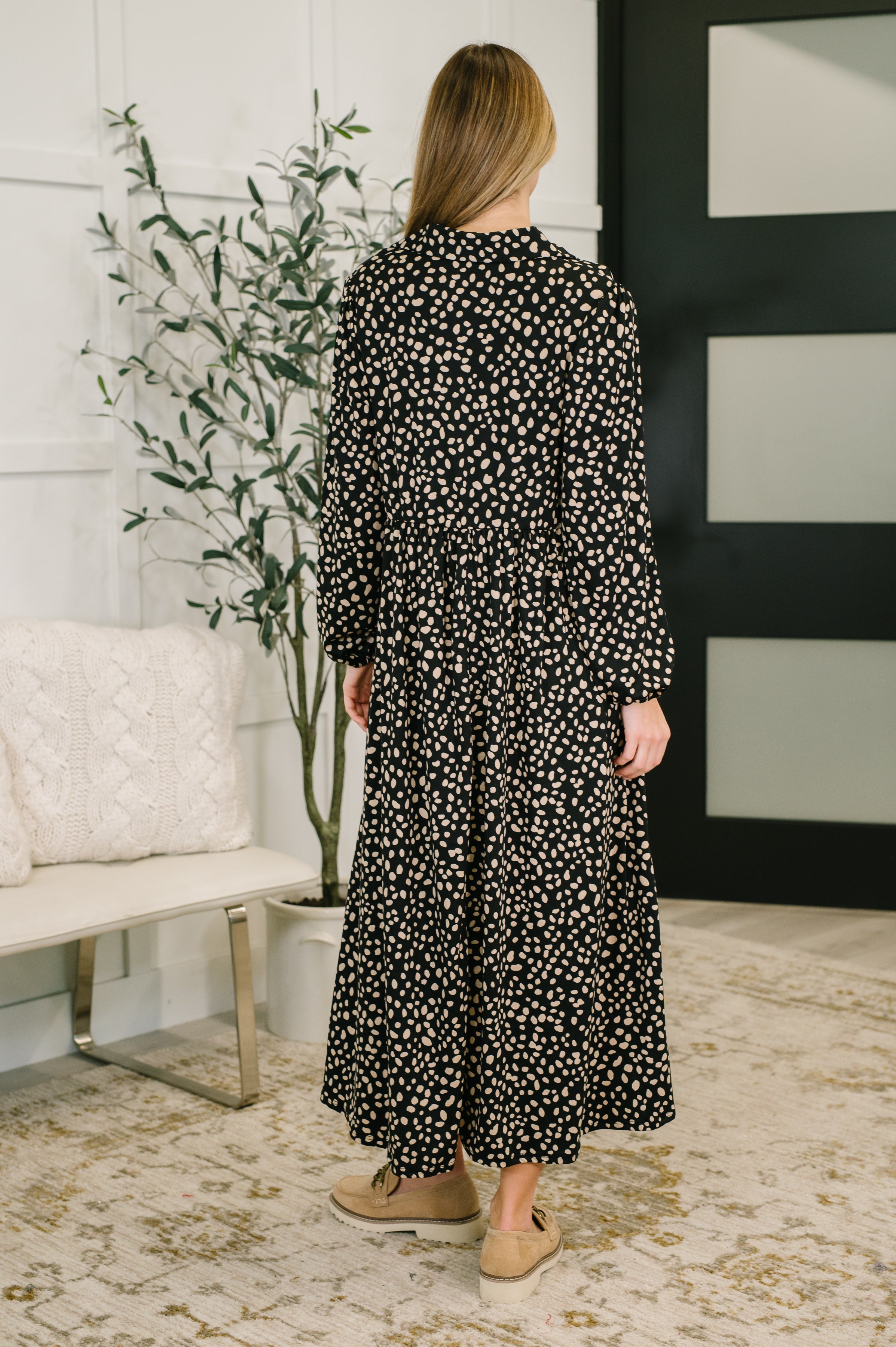 Woman wearing a black collared maxi dress with brown spots and tan shoes in an indoor setting.