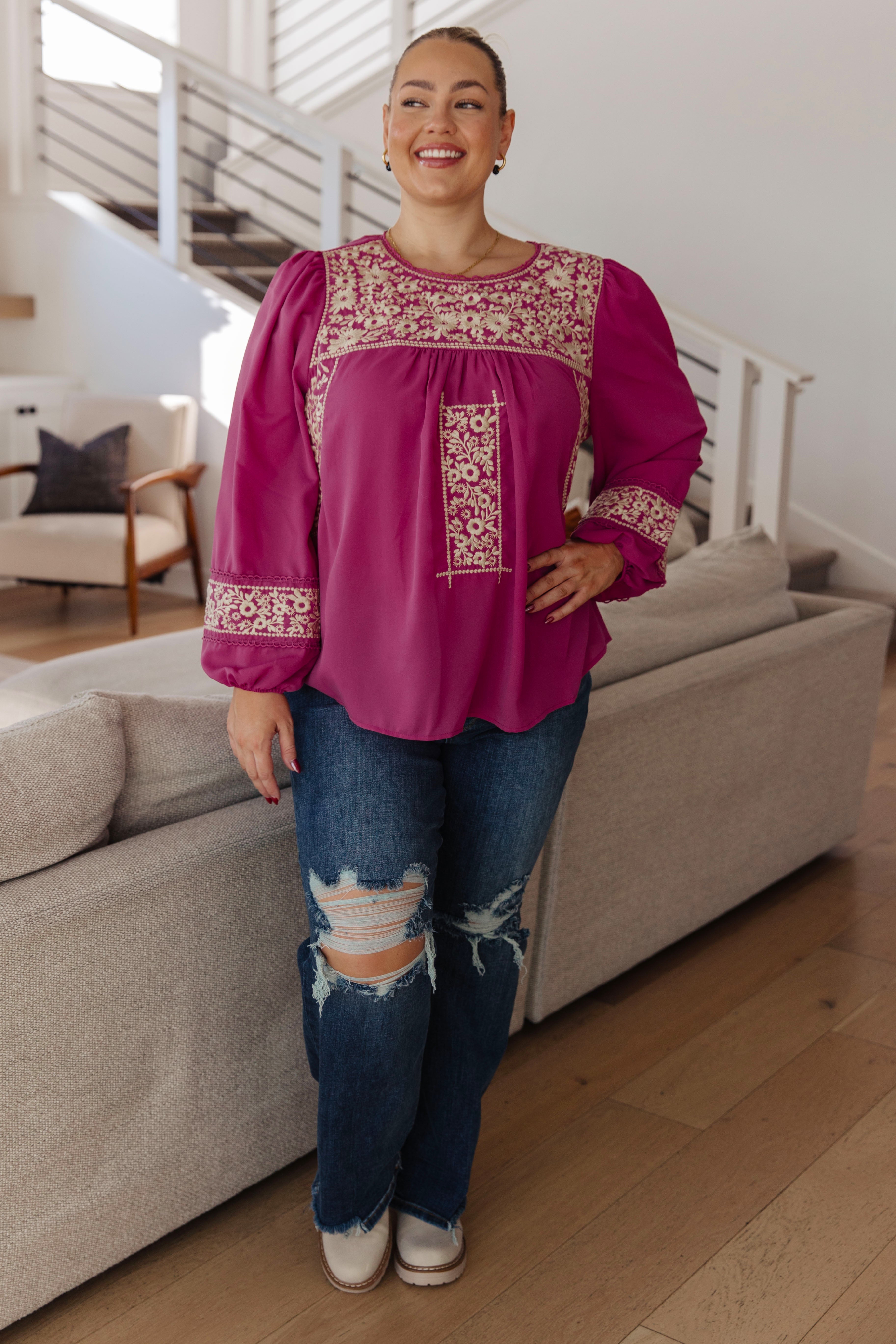 Woman wearing a magenta long-sleeve blouse with intricate white embroidery, distressed blue jeans and black and white sneakers standing in an indoor setting.