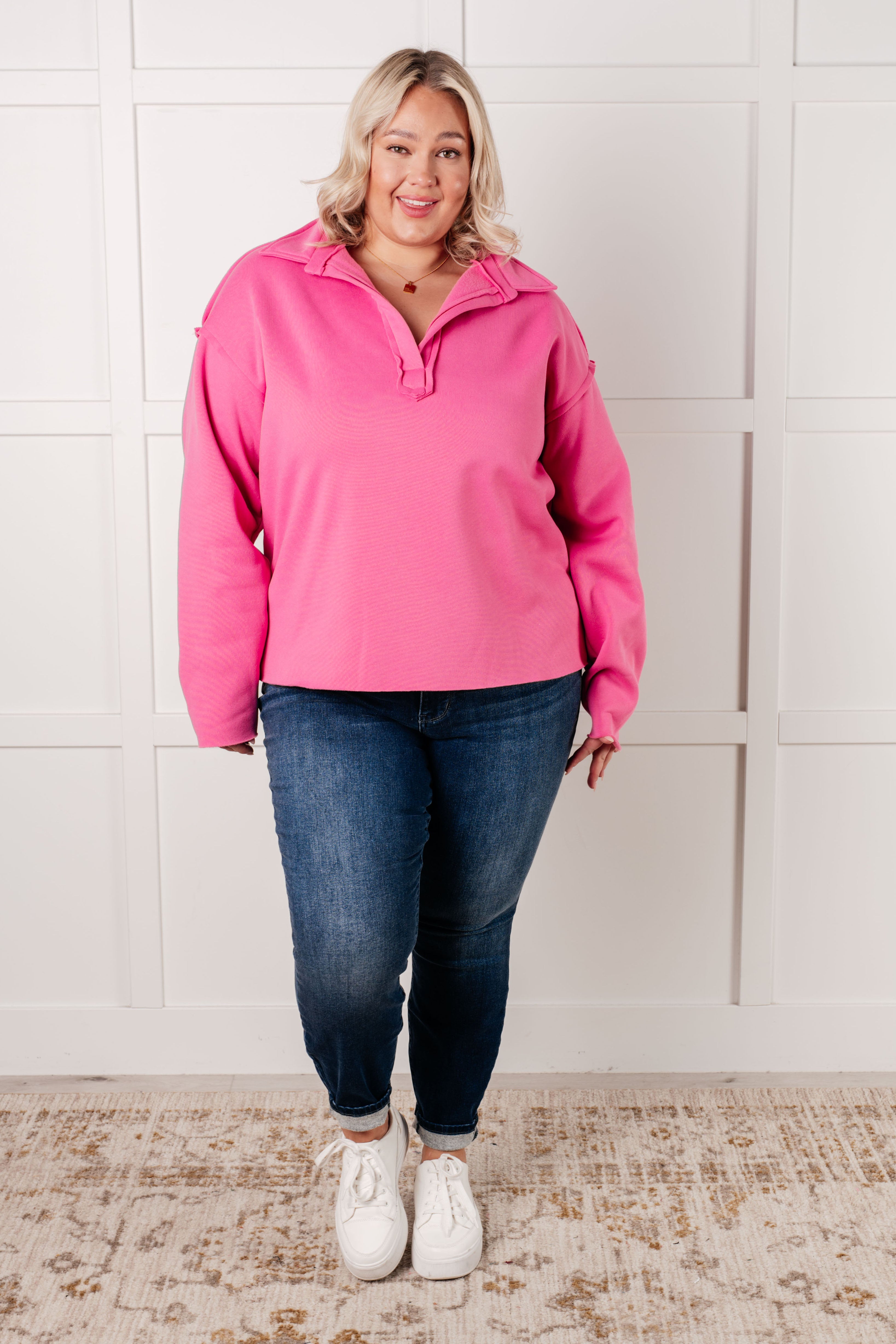 Woman wearing a hot pink colored pullover with exposed seams, blue jeans and white sneakers with a white background.