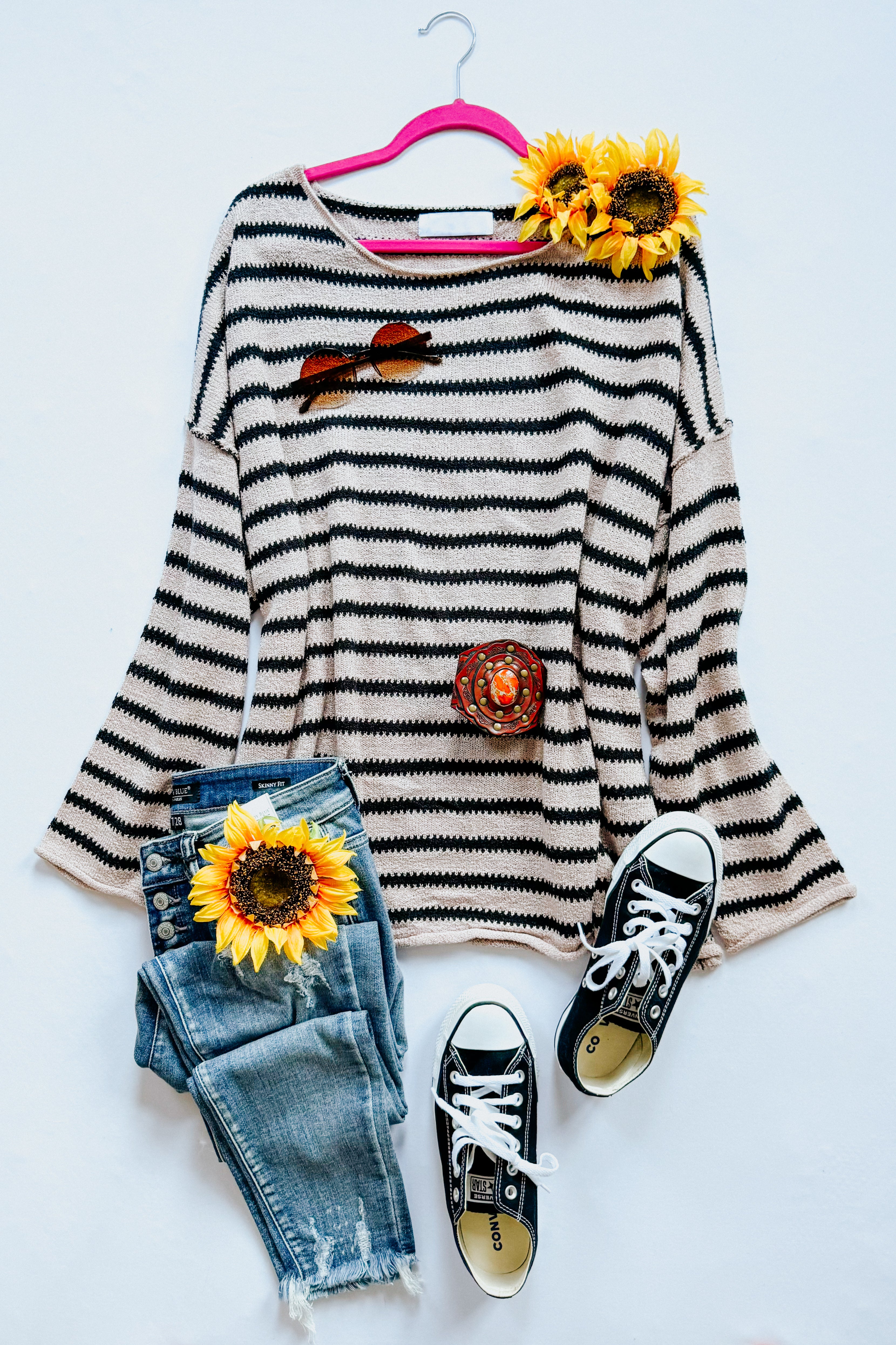 Flat lay on a white background with a taupe long sleeve sweater with black stripes, blue jeans, black converse sneakers, accessories and floral elements.