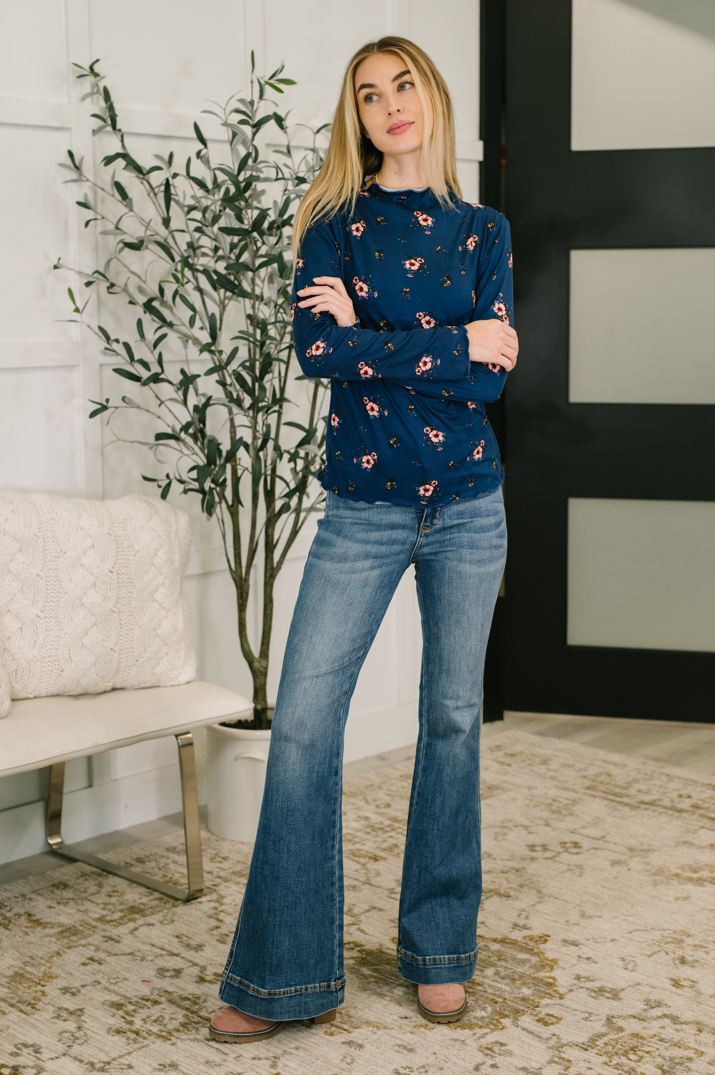 Woman wearing a blue floral top, blue flared jeans and brown boots standing in a room with a plant and bench.