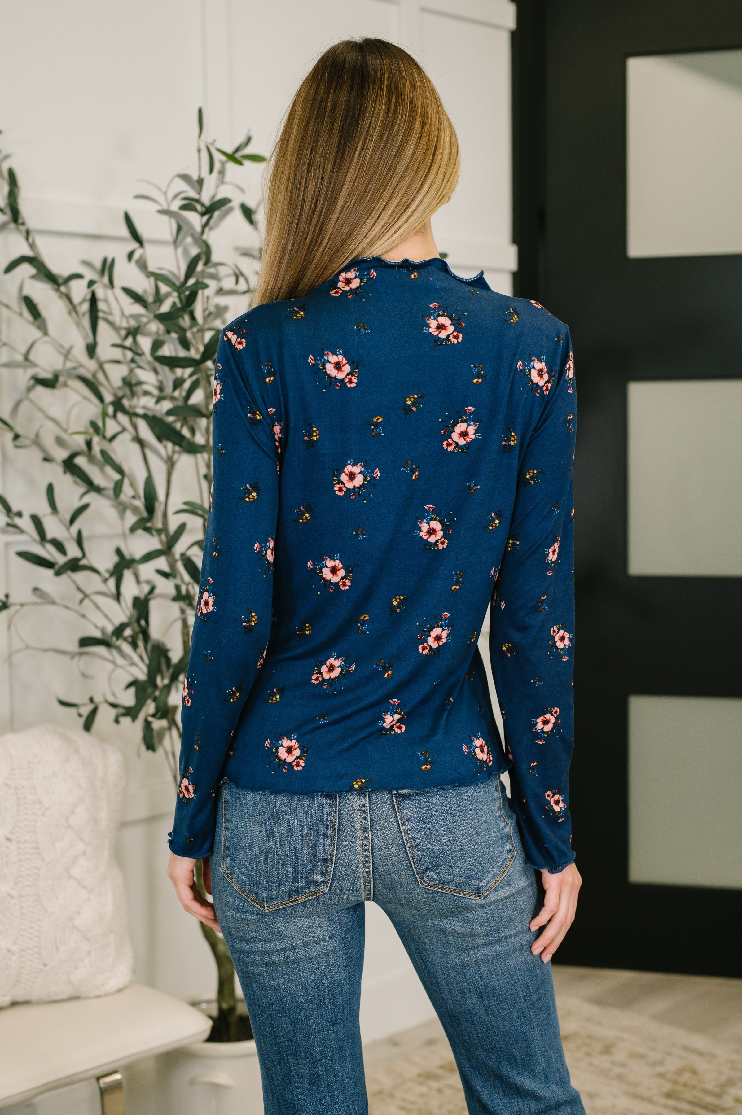 Woman with her back facing the camera wearing a blue floral top and blue jeans in an indoor setting.