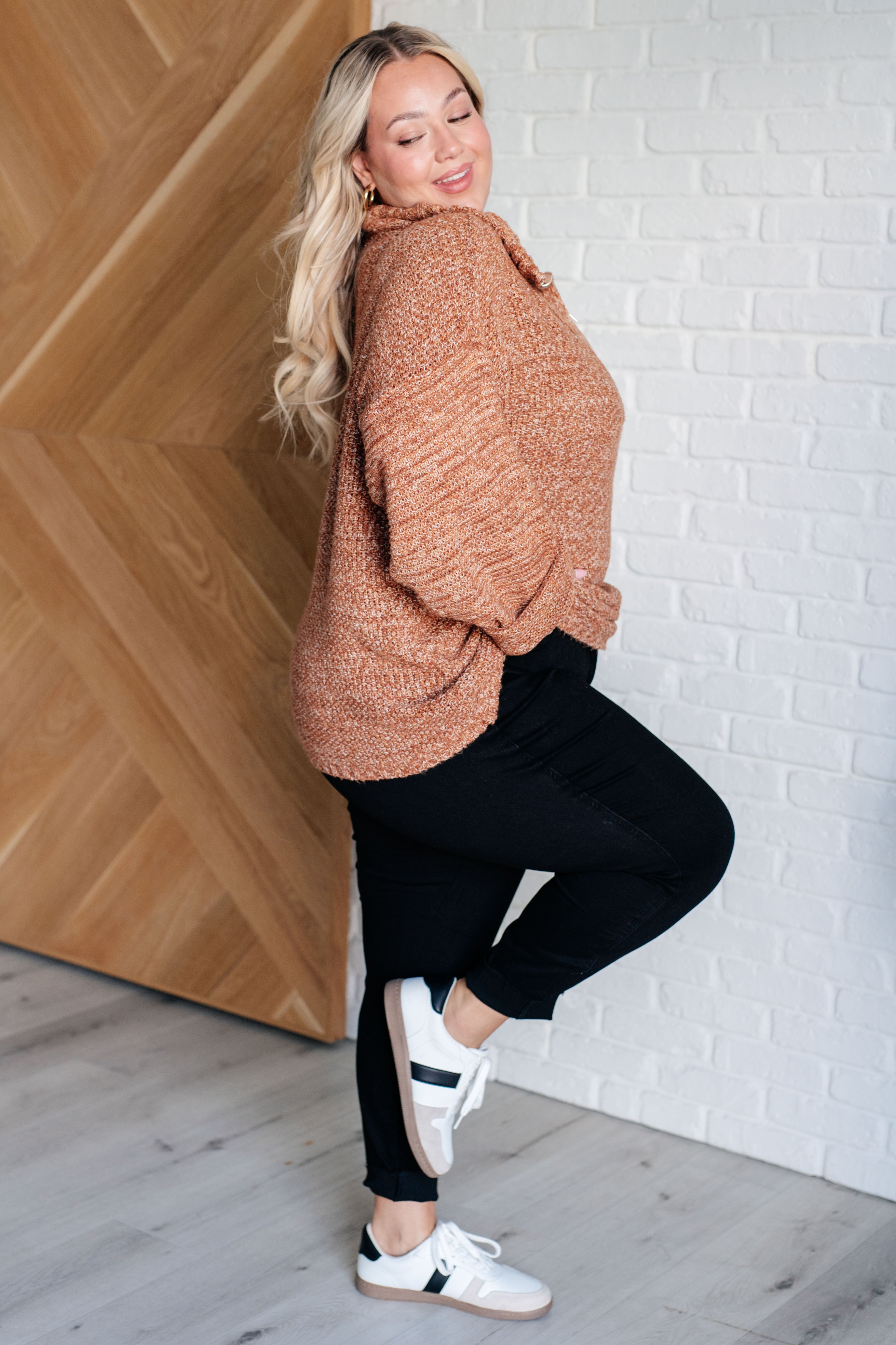 Woman wearing a rust colored oversized long-sleeve sweater, black leggings and white sneakers standing against a white brick wall.