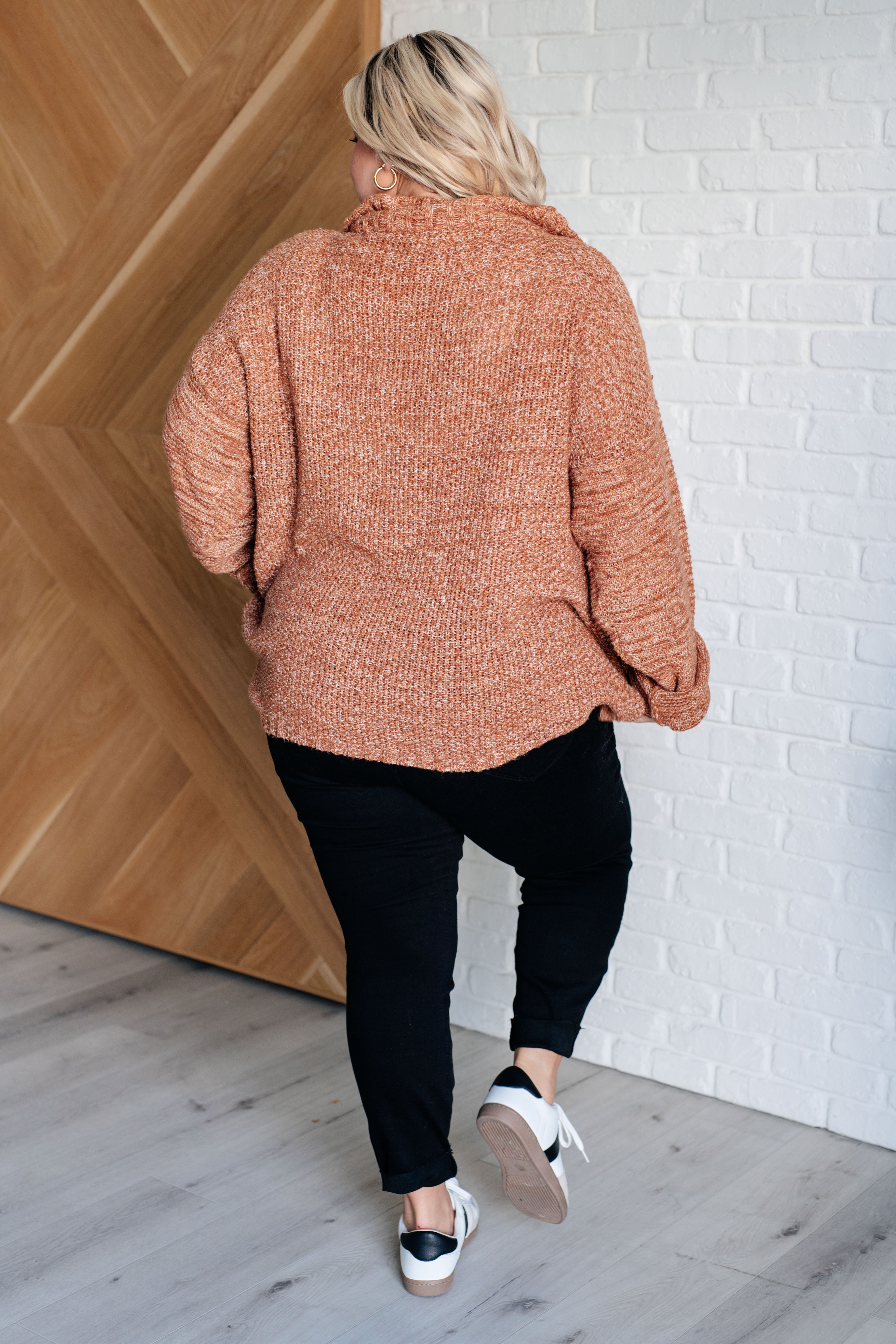 Woman wearing a rust colored oversized long-sleeve sweater, black leggings and white sneakers standing against a white brick wall.