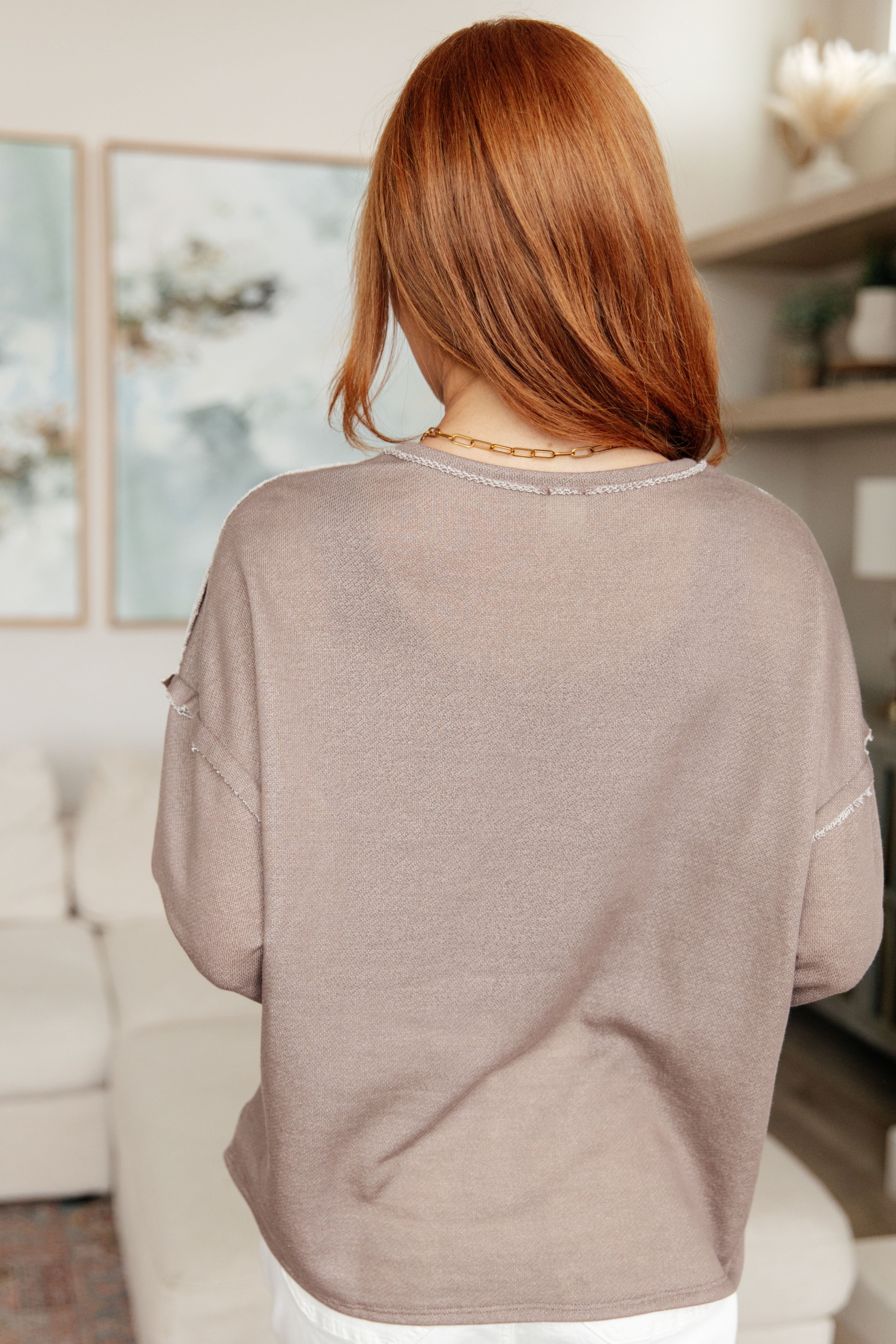 Woman wearing a taupe reverse stitch v-neck pullover with raw accents and light beige pants standing in an indoor setting.