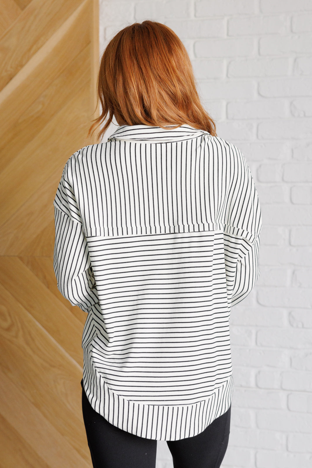 Woman wearing a white and black striped collared pullover top and black leggings standing against a white brick wall.