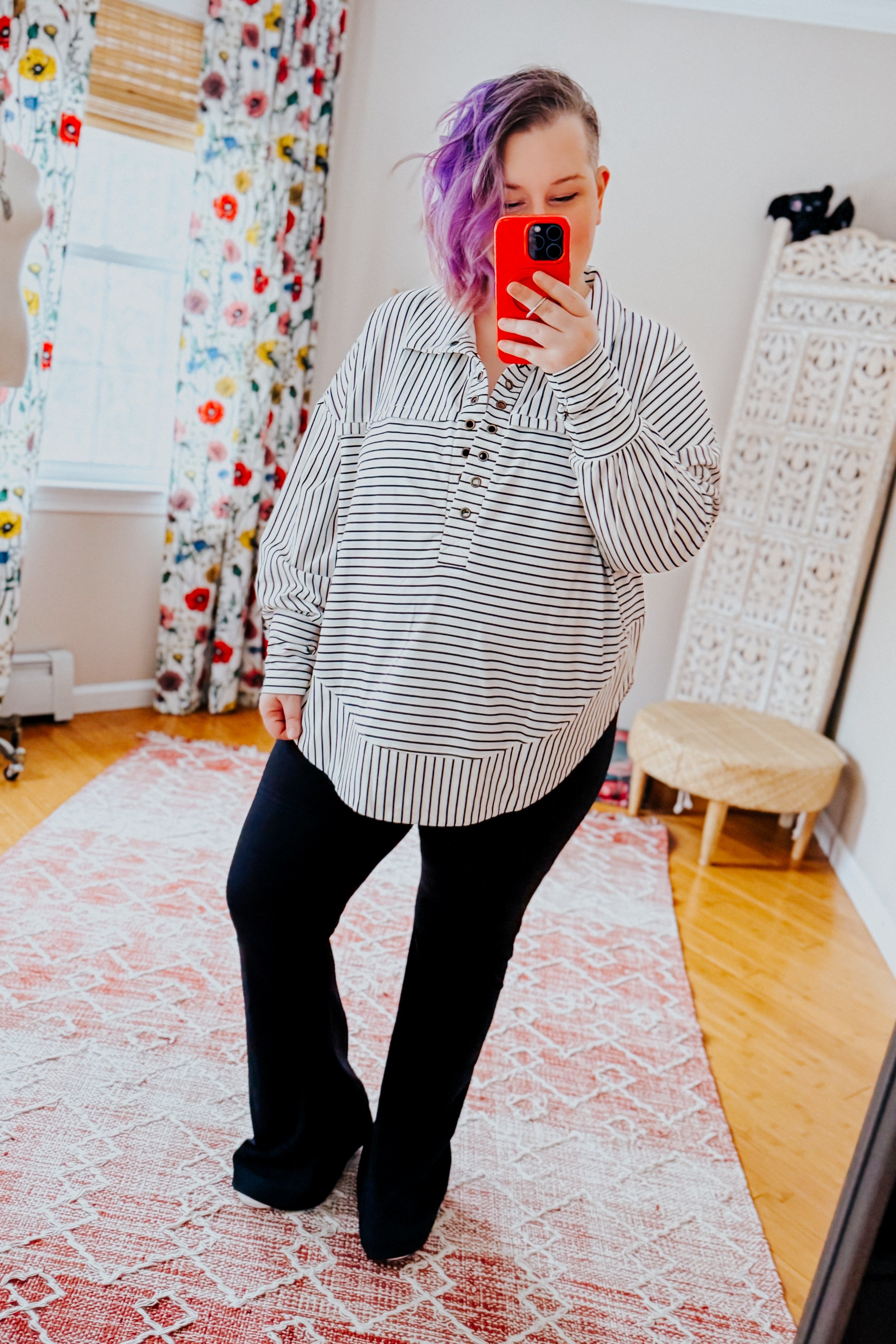 A woman taking a mirror selfie wearing a white and black striped collared pullover top, black pants and black converse sneakers standing in an indoor setting.