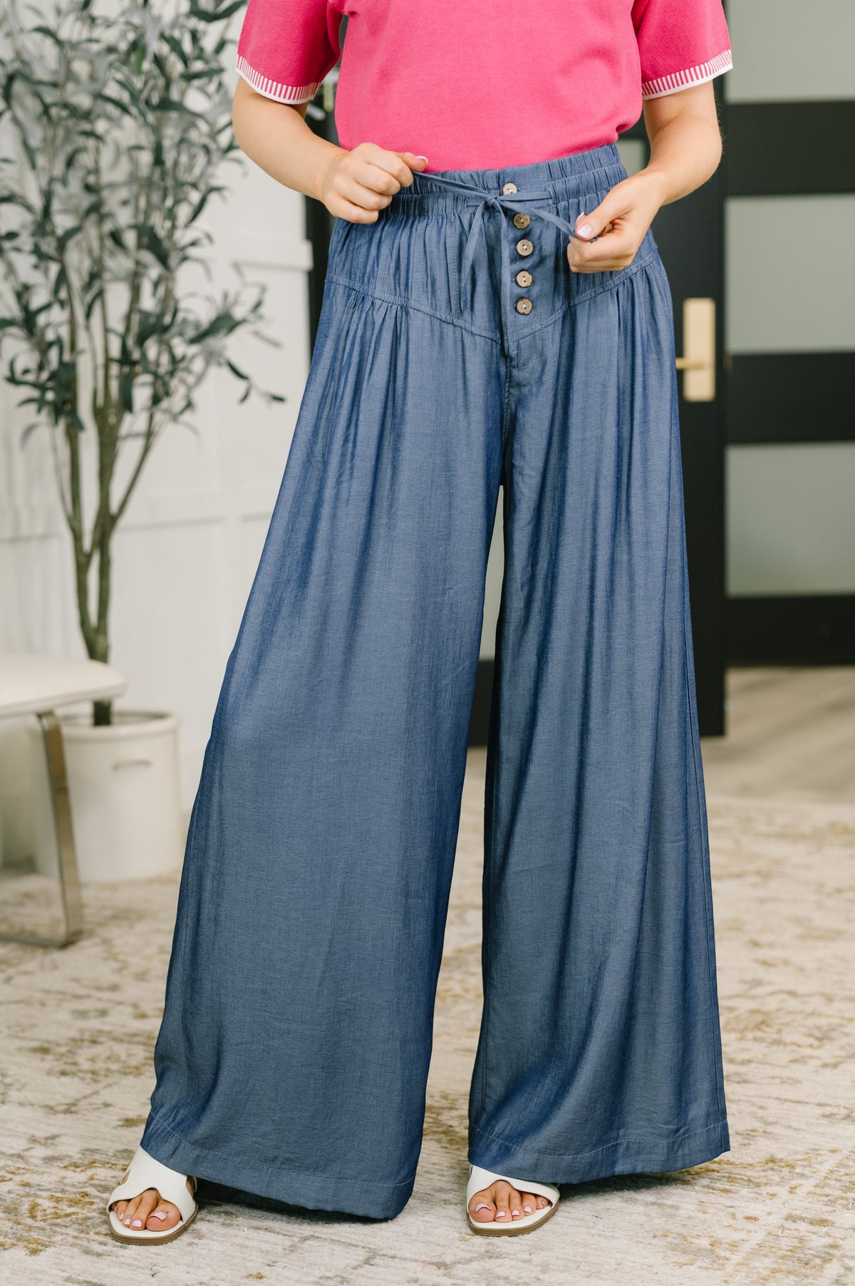 Woman wearing blue high waisted wide leg bottoms with front buttons and a pink top indoors with sandals