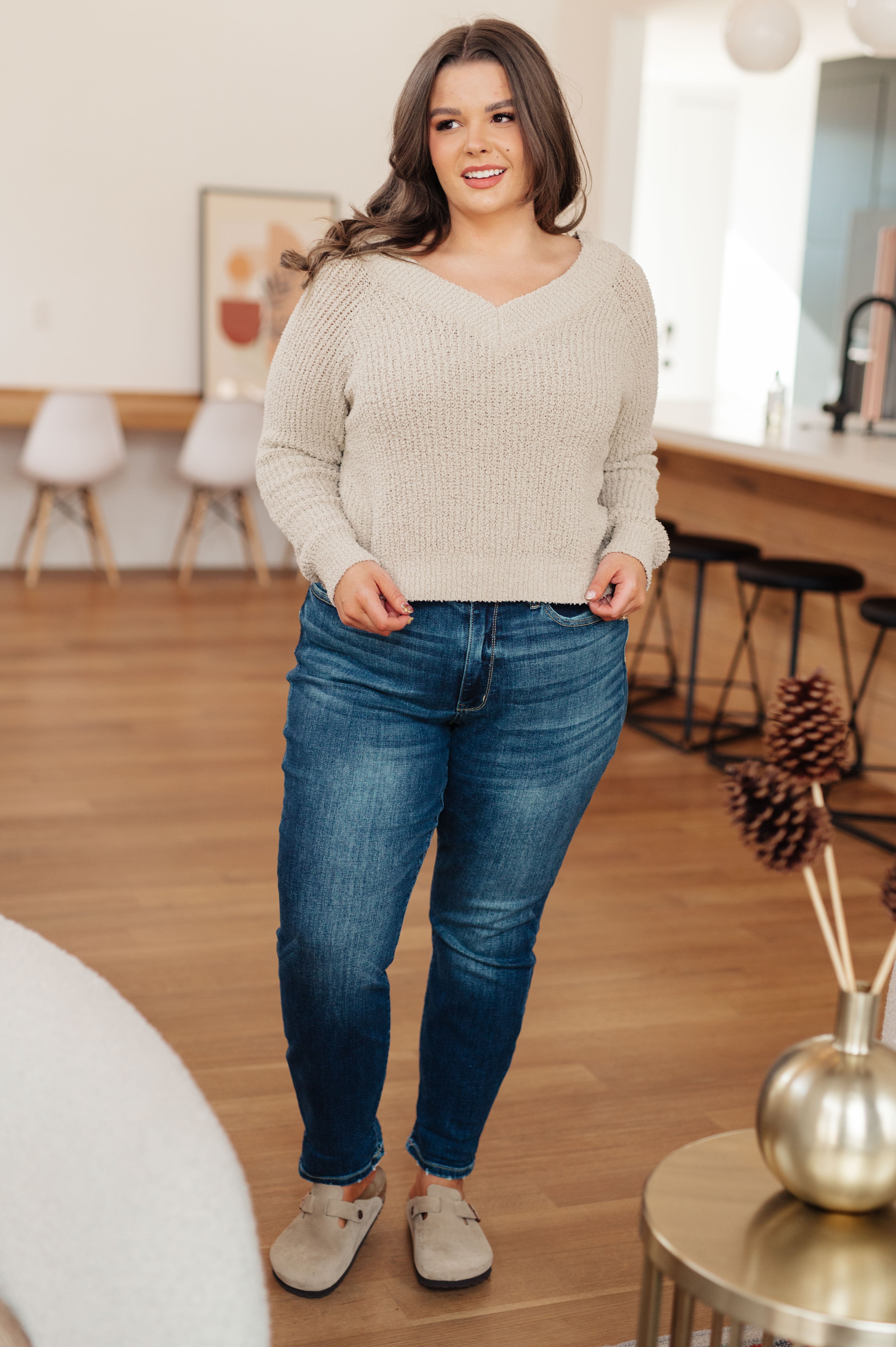 Woman wearing a beige sweater, blue jeans and beige mules standing in a room with wooden flooring and decor.