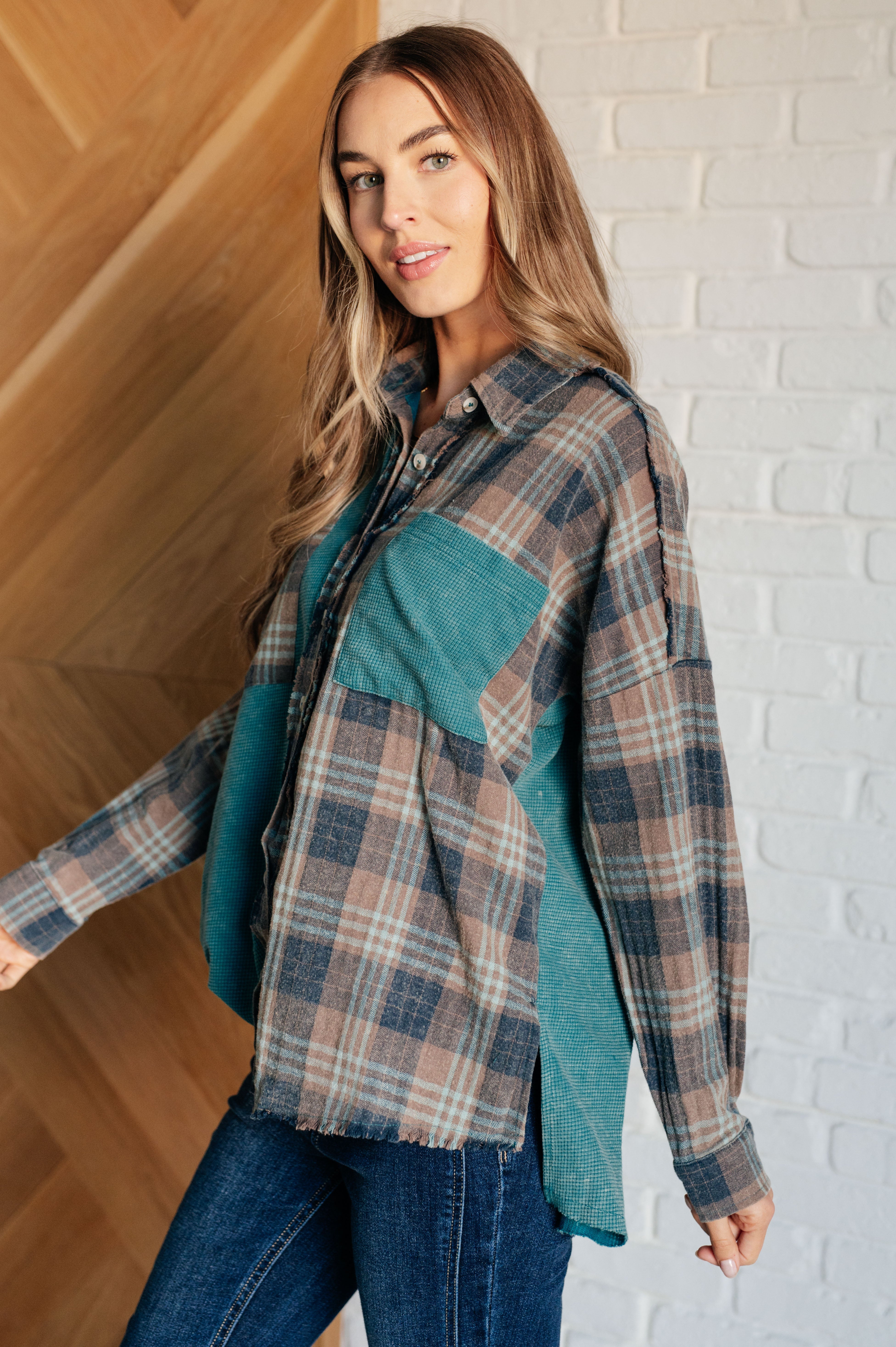 Woman wearing a blue plaid button up shirt over blue jeans against a white brick wall.