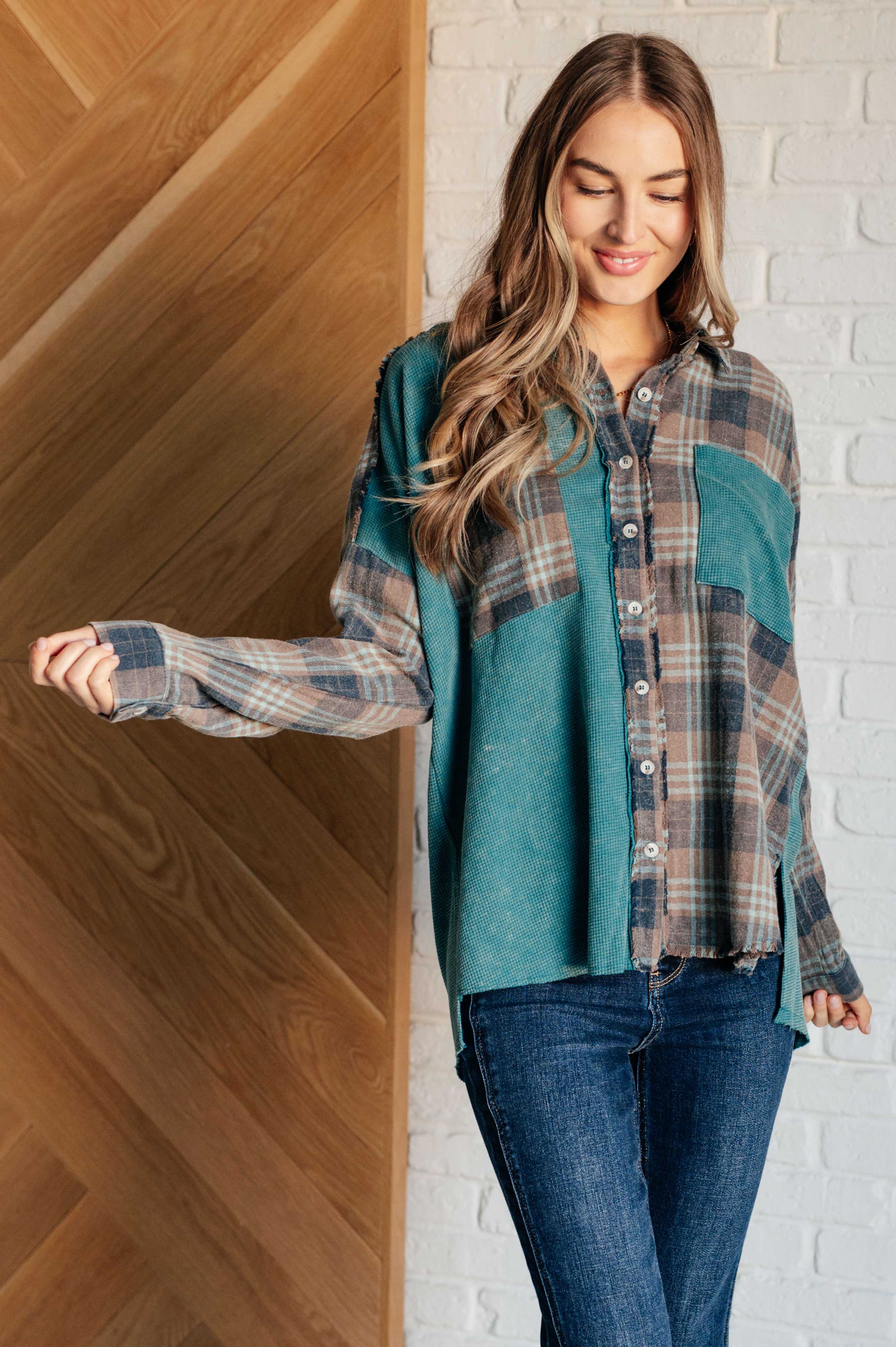 Woman wearing a blue plaid button up shirt over a jeans, standing against a wooden and brick wall.