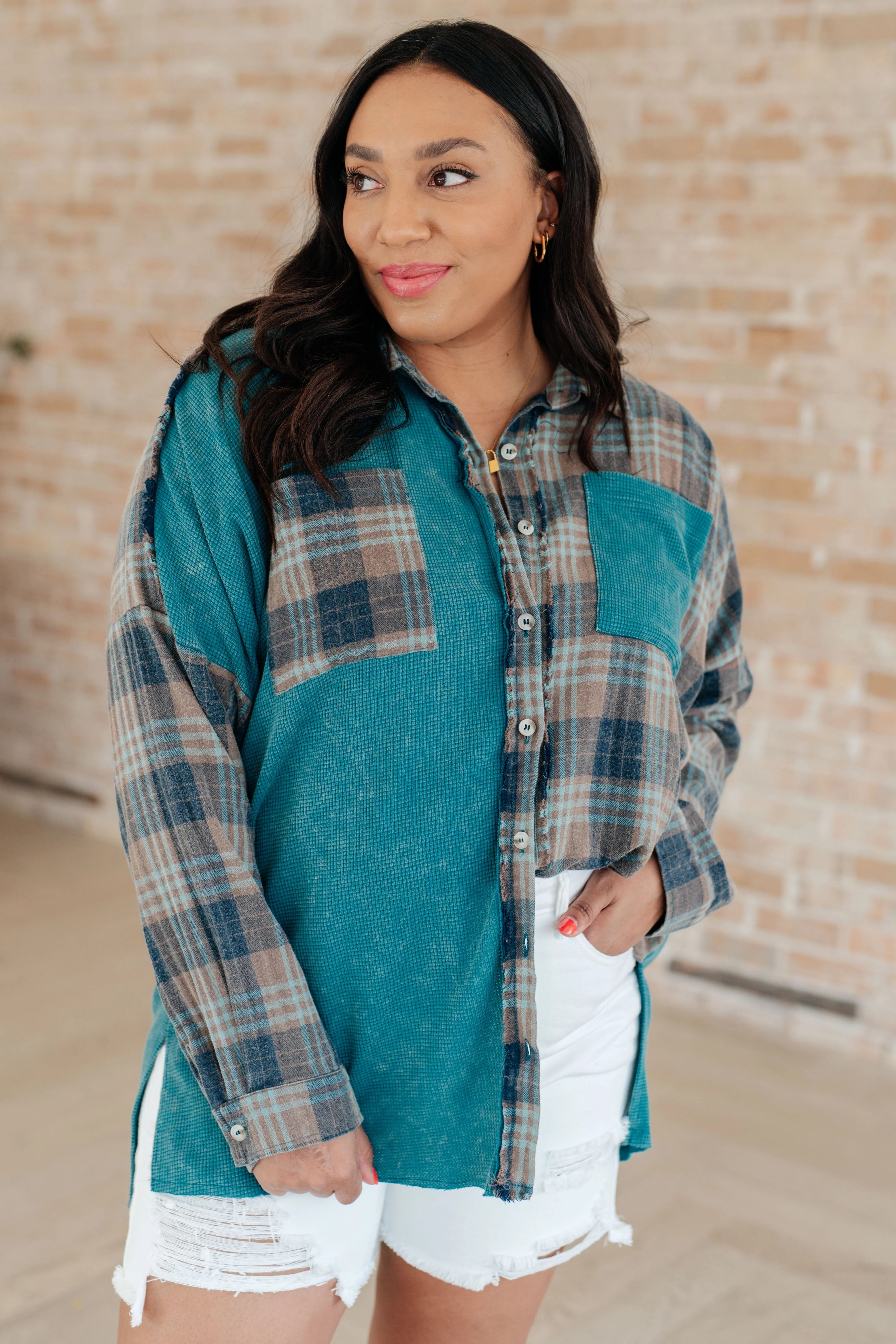 Woman wearing a blue plaid button up shirt with white distressed shorts against a brick wall.