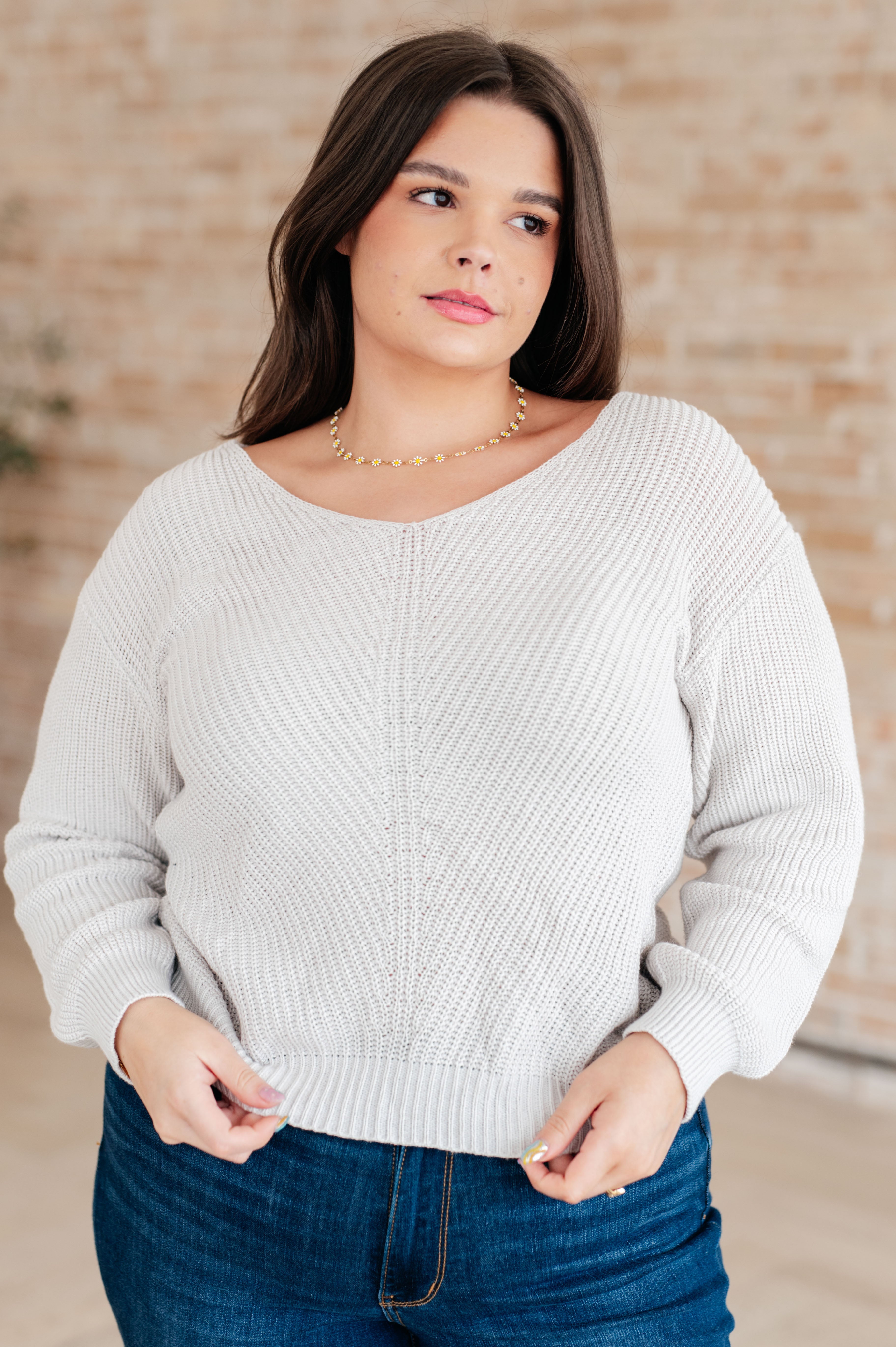 Woman wearing a light grey knit v-neck sweater and blue jeans standing against a brick wall.