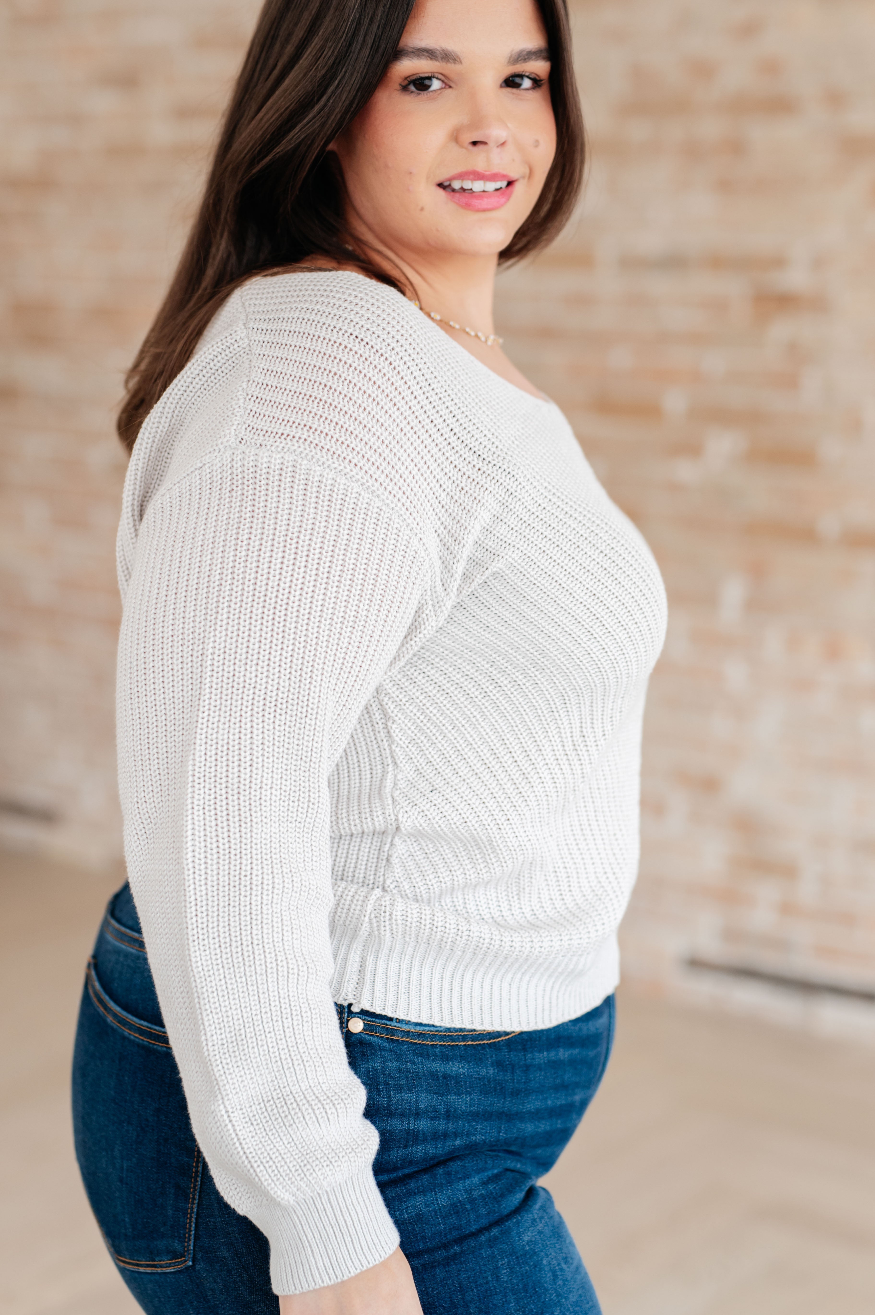 Woman wearing a light grey knit v-neck sweater and blue jeans standing against a brick wall.