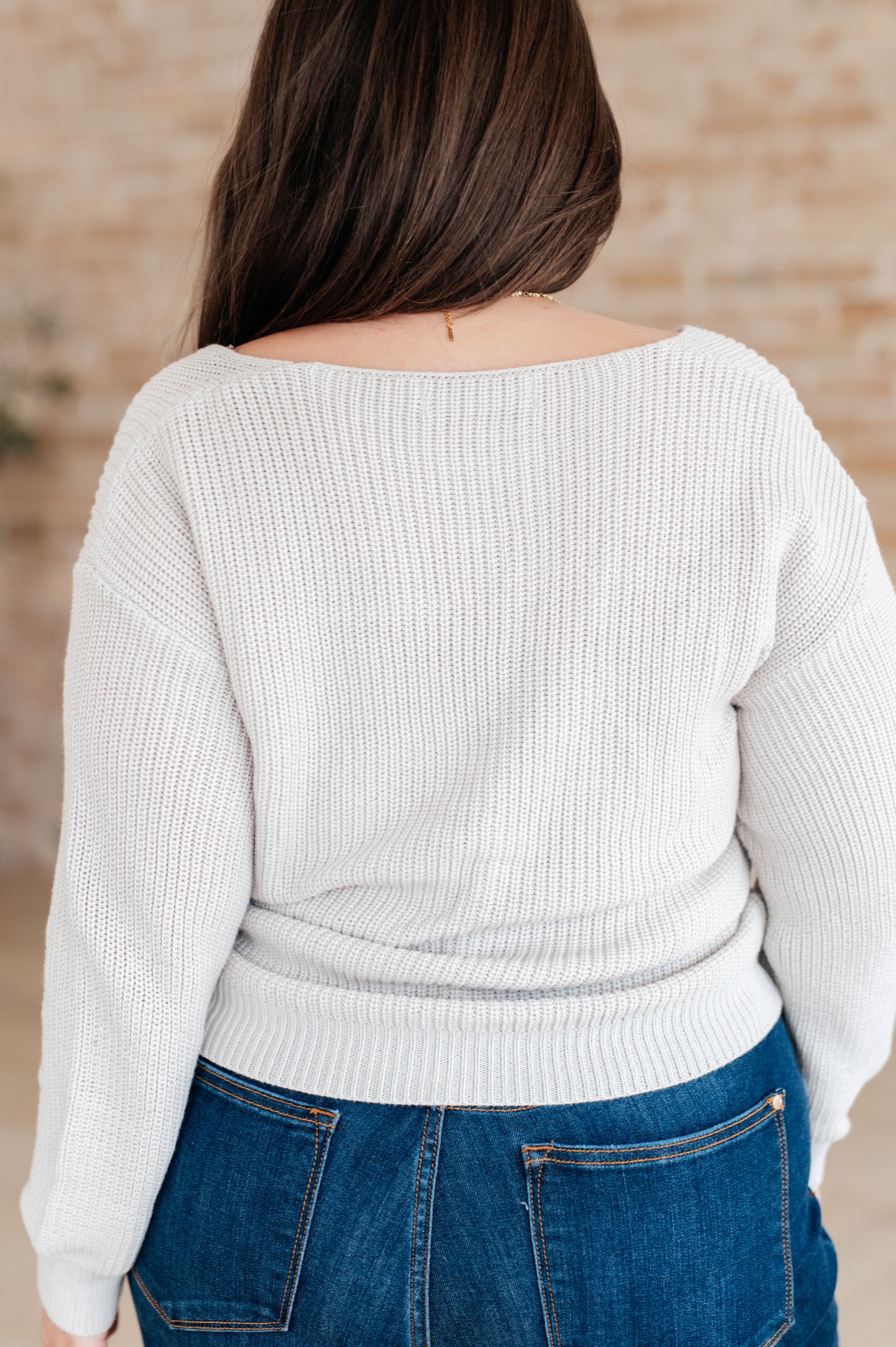 Woman wearing a light grey knit v-neck sweater and blue jeans standing against a brick wall.