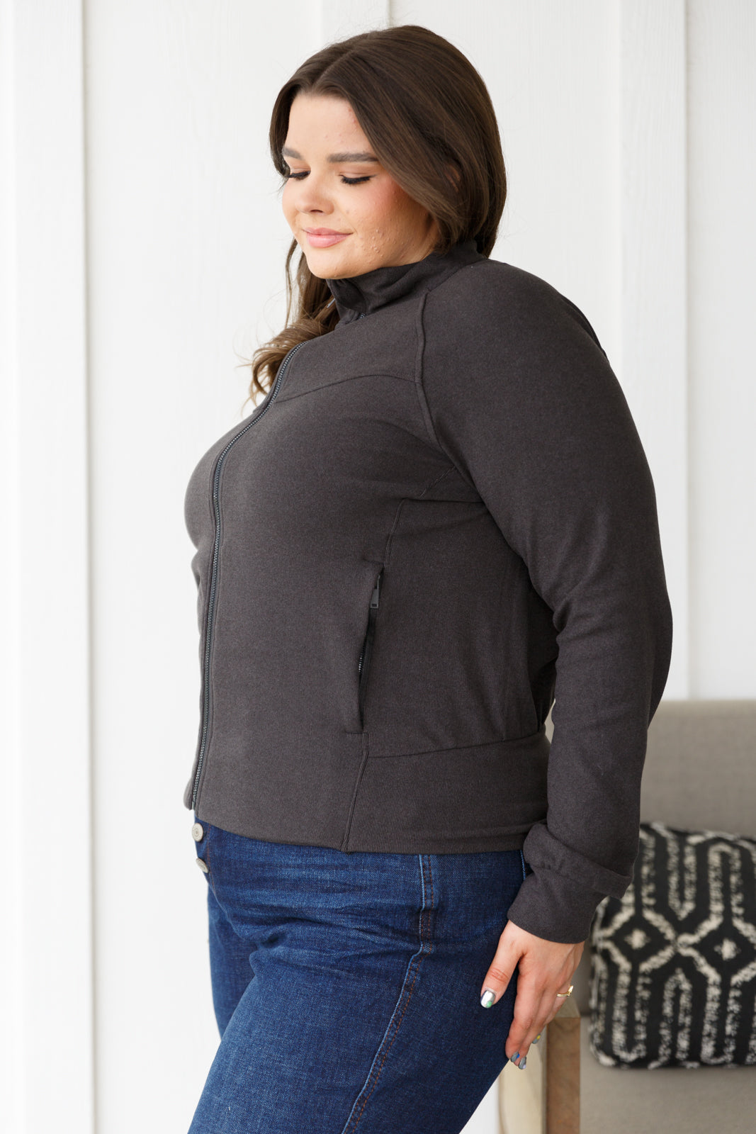 Woman wearing a dark gray jacket and blue jeans standing against a white wall.