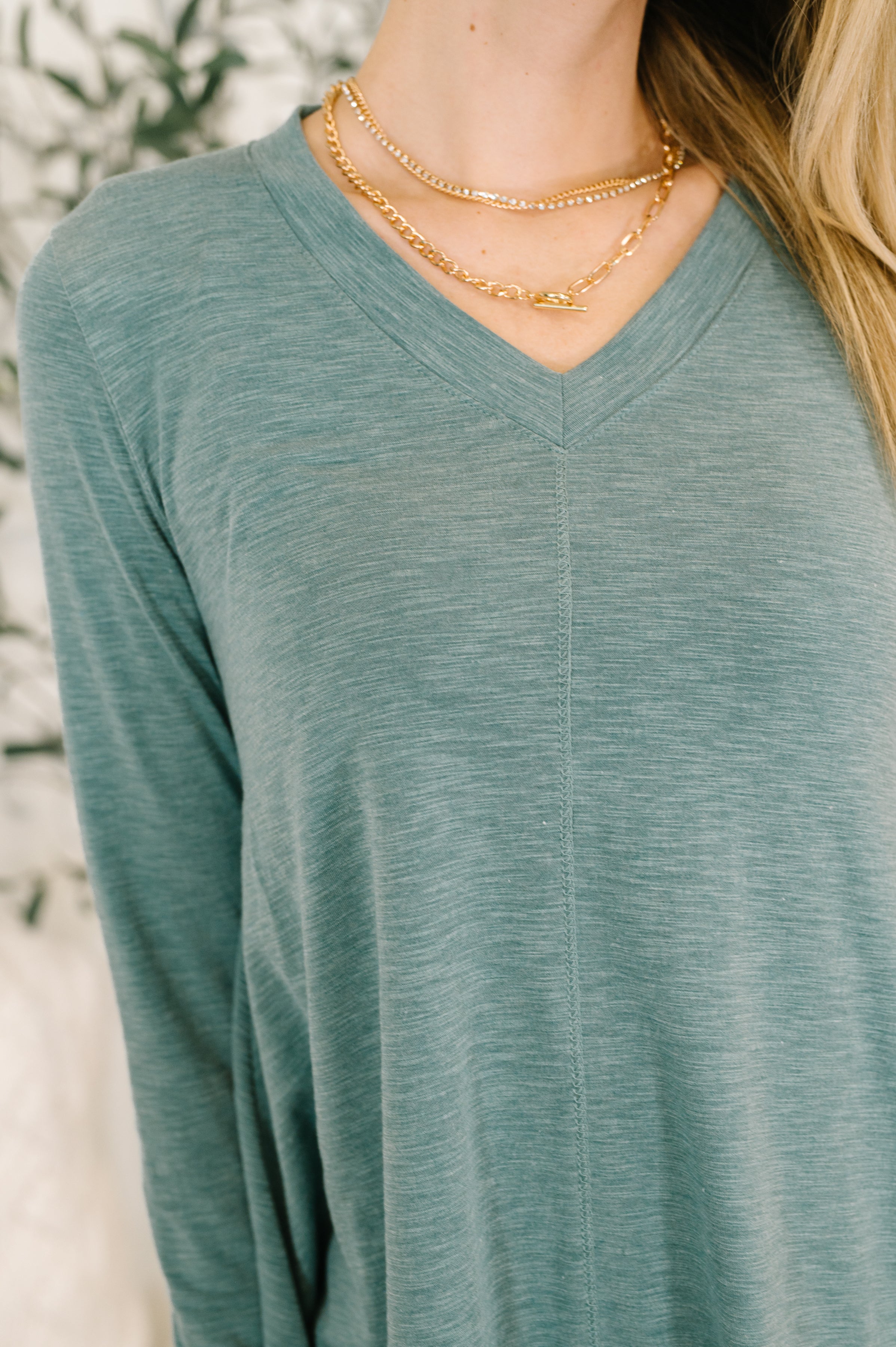 Close up shot of a woman wearing a light teal v neck long sleeve top and a gold necklace with a blurred background.